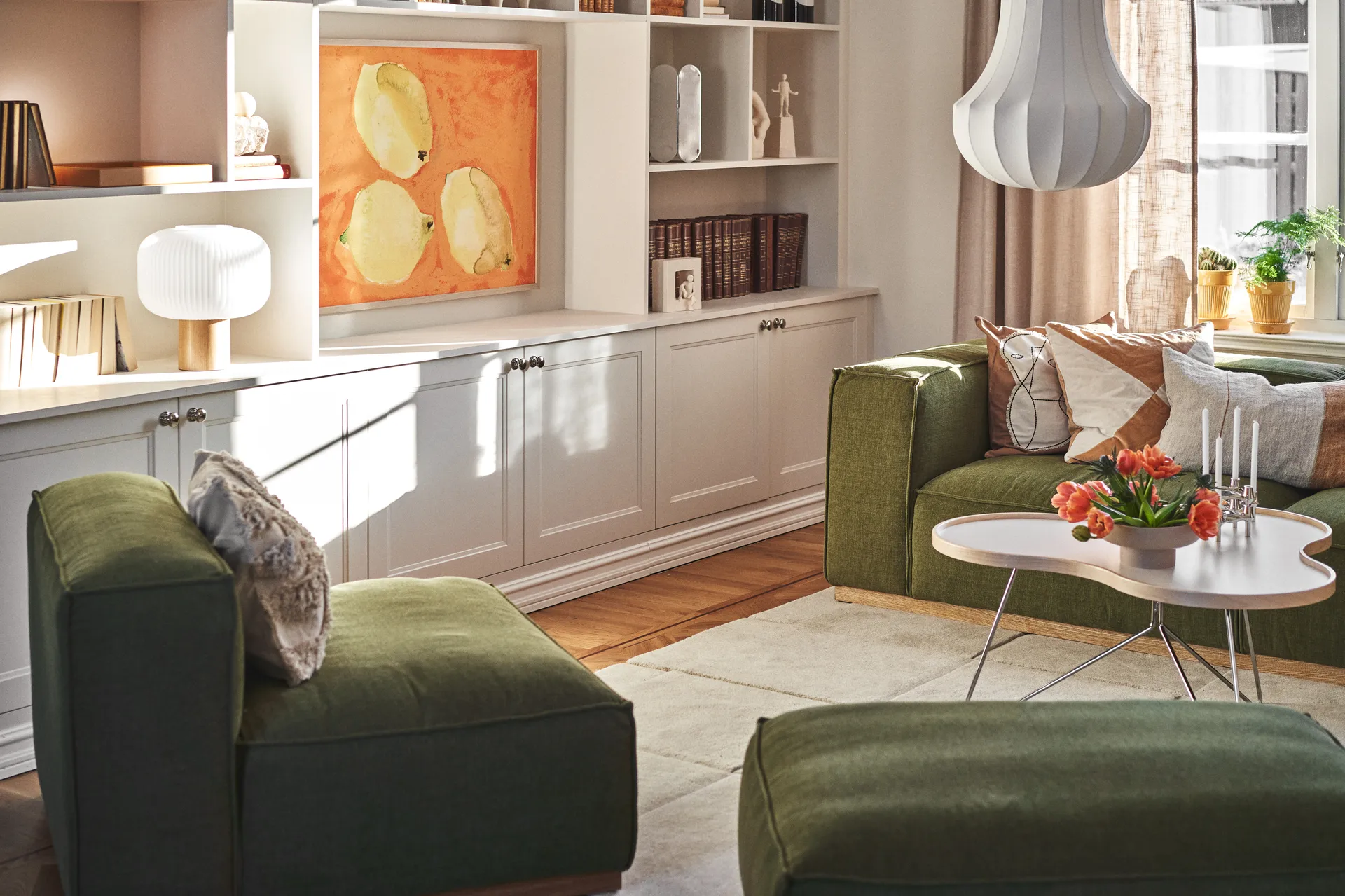 Solrigt stue med grønne sofaer, et hvidt sofabord med orange tulipaner og lys, samt indbyggede hvide hylder med kunst.