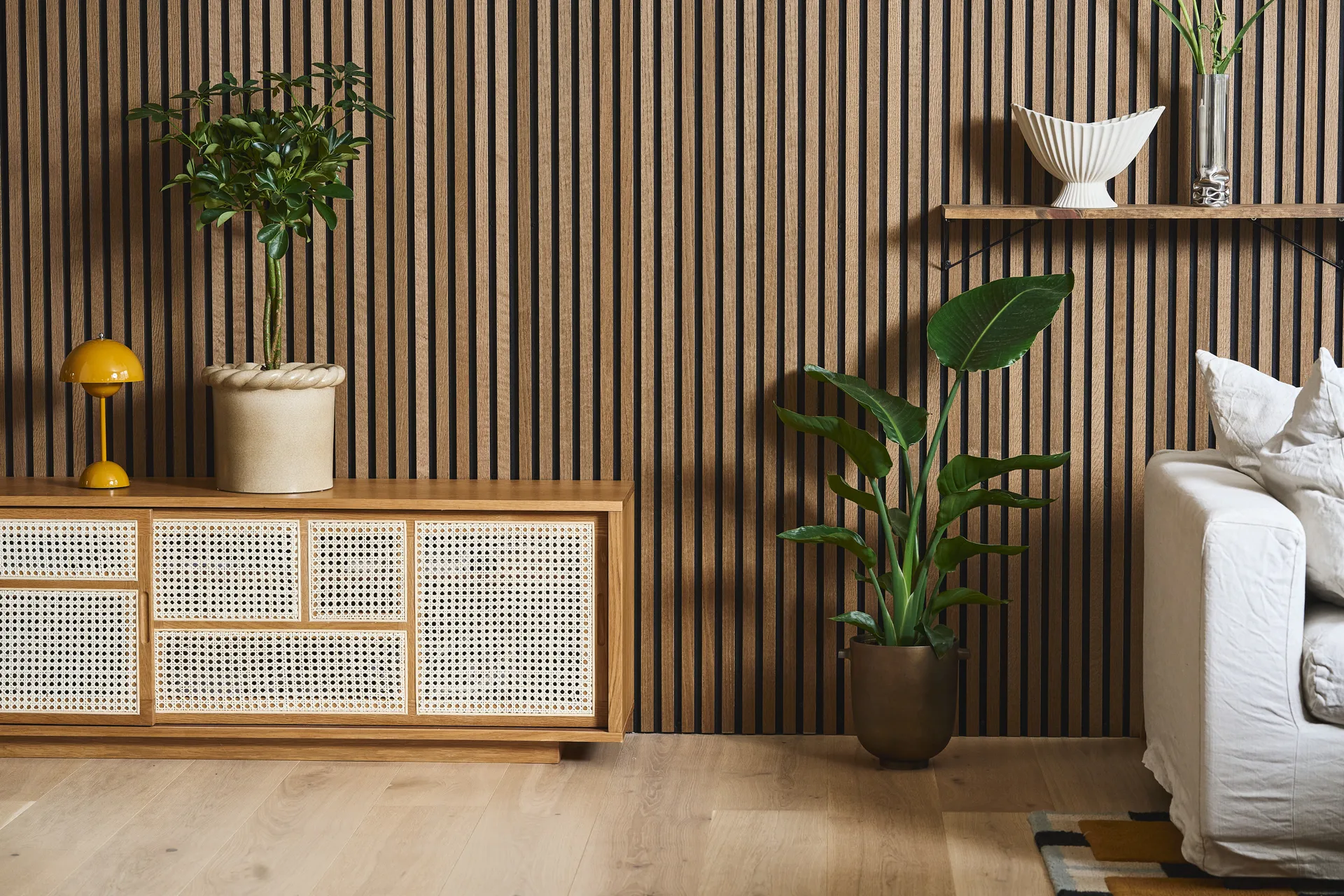 Moderne stue med lameltrævæg, rottingmøbel, gul lampe, forskellige potteplanter og en hvid sofa.