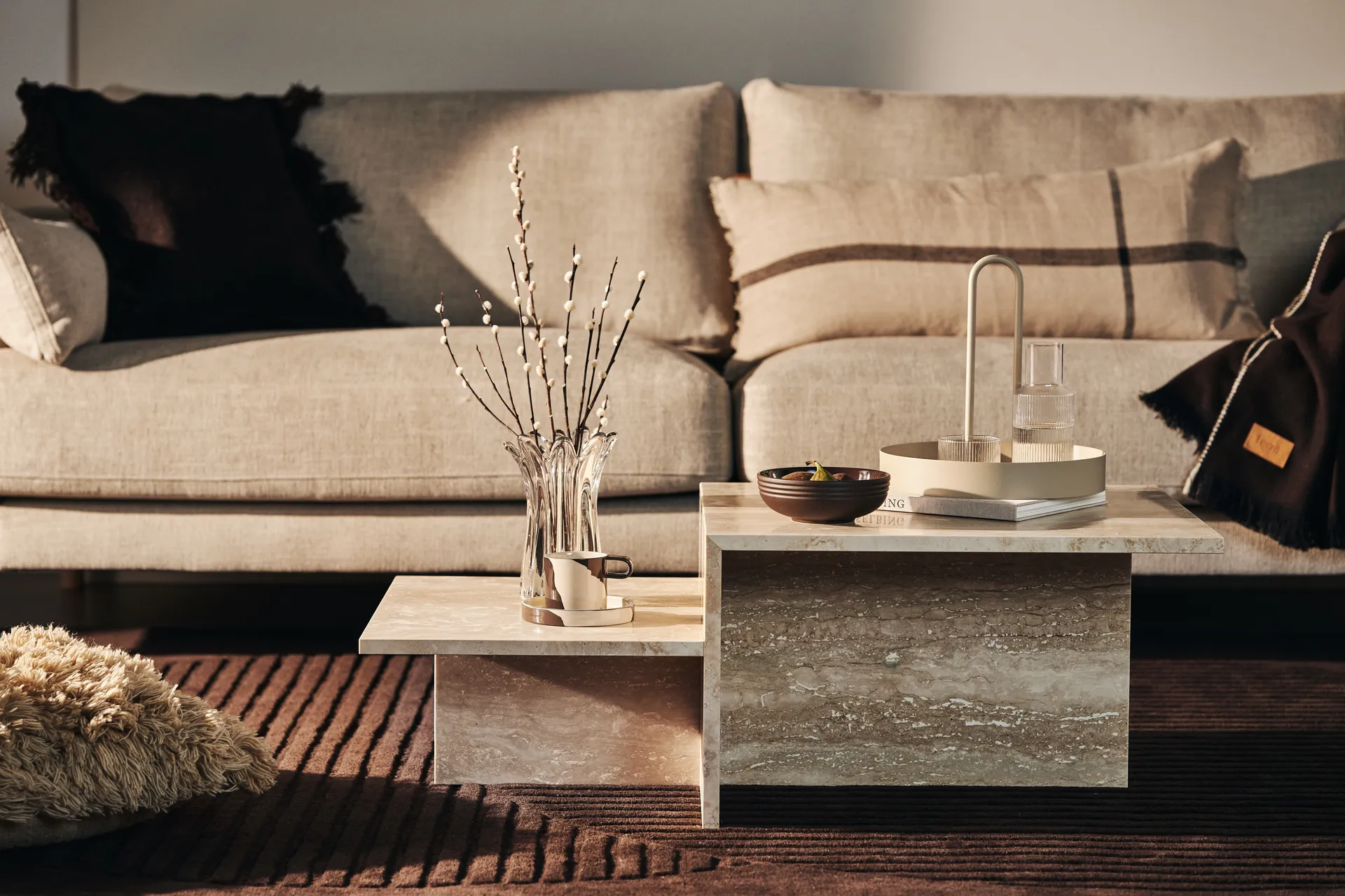 Niveaudelt sofabord i beige sten foran en neutral sofa med dekorativt tilbehør.