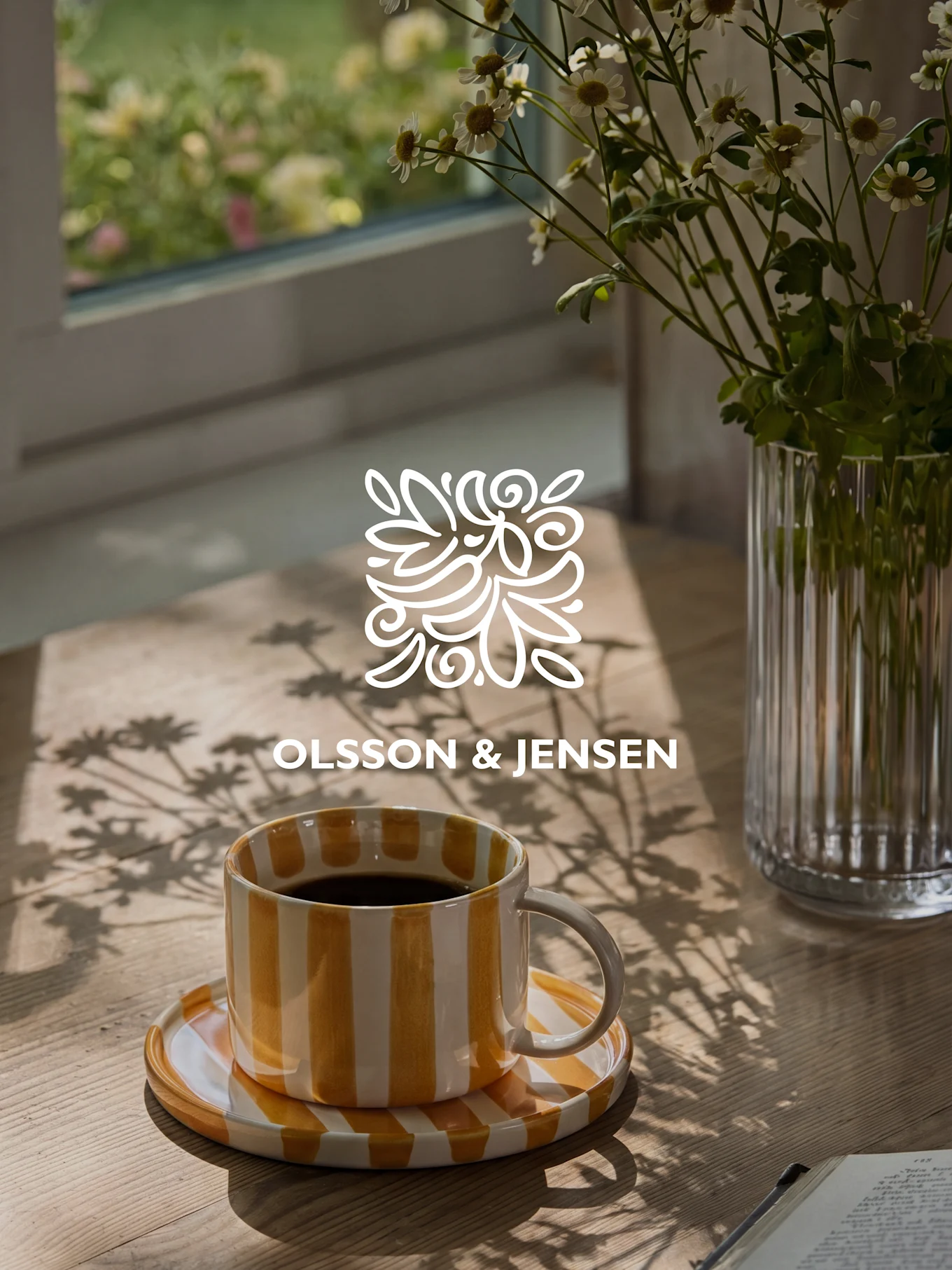 Gul- og hvidstribet kaffekop på et træbord med en vase med hvide blomster og solbeskinnede skygger, Olsson & Jensen logo.