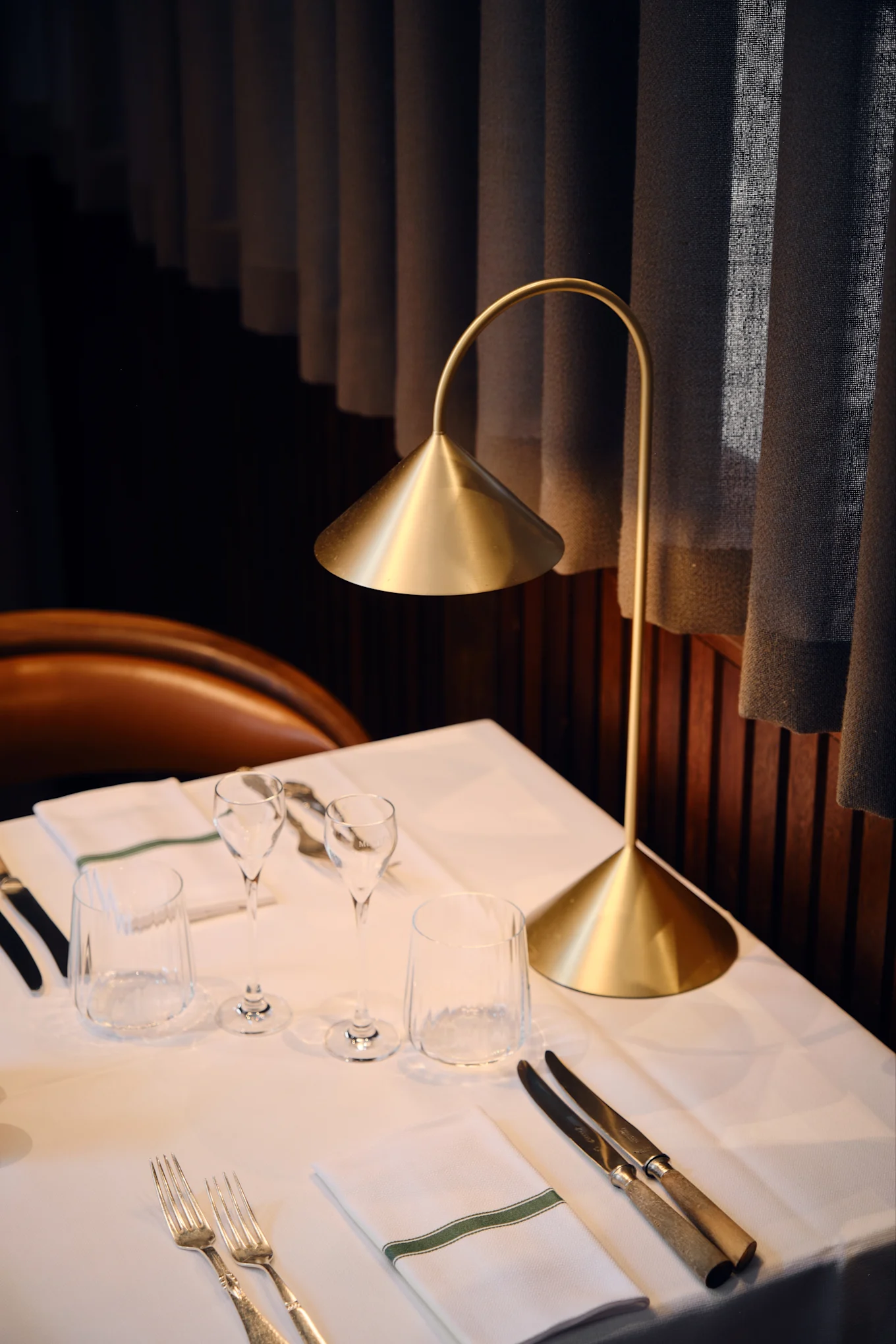En moderne guldbordslampe oplyser et dækket restaurantbord med hvidt linned, glas, bestik og en serviet med grøn stribe.