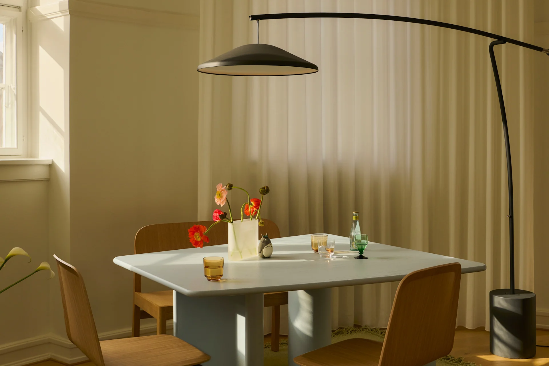 En moderne spisestue med et lyseblåt bord, træstole, en sort buet gulvlampe og en vase med farverige blomster i sollys.