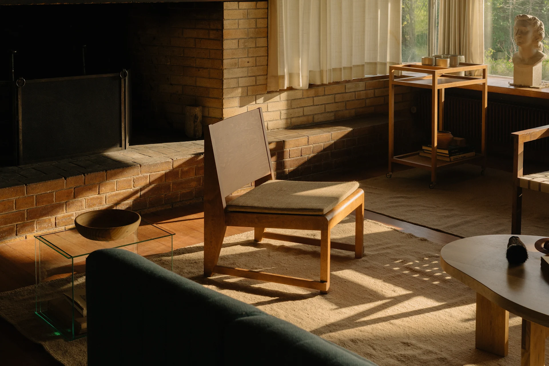 En solrig stue med pejs af mursten, en træstol og en grøn sofa, med en busteskulptur ved vinduet.