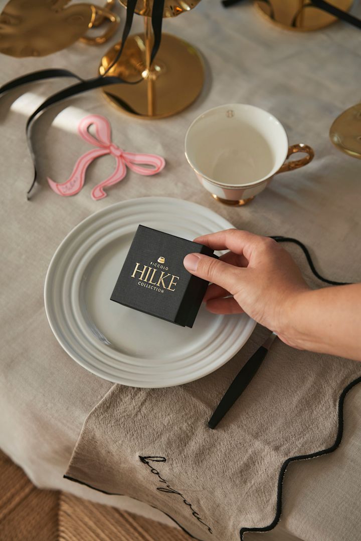 Fejr Galentine's Day hjemme med dine bedste veninder. Her kan du se en hånd der lægger en boks fra Hilke Collection ved en af gæsternes plads som en lille gave.