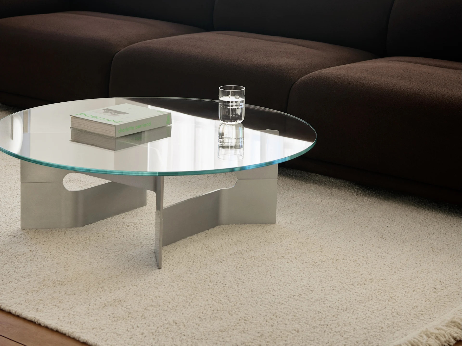 Rundt glas sofabord med bøger og et glas vand på et cremefarvet, loddent tæppe foran en mørkebrun sofa.