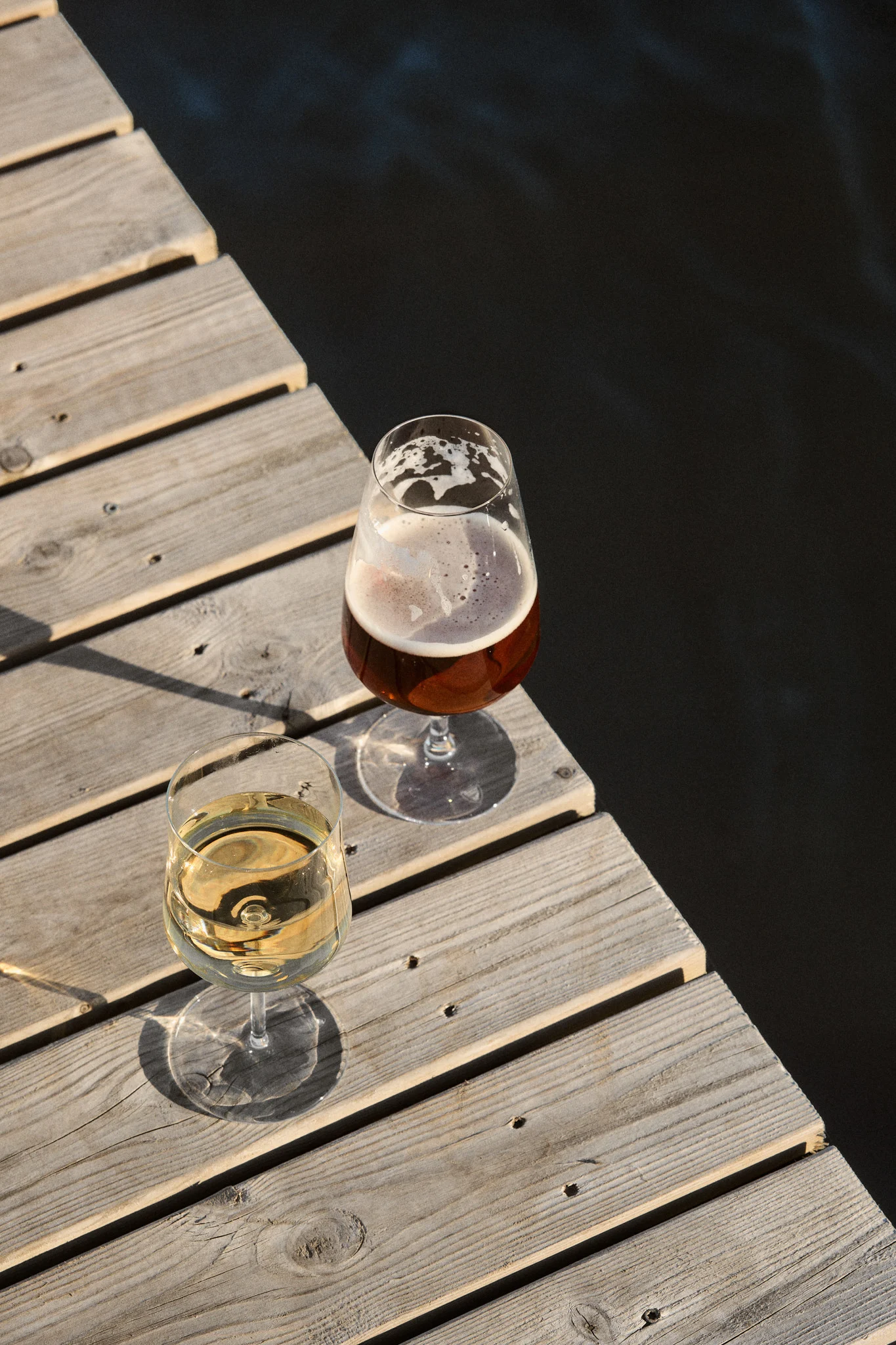 To glas, et med hvidvin og et med øl, på en træbro ved mørkt vand i sollys.