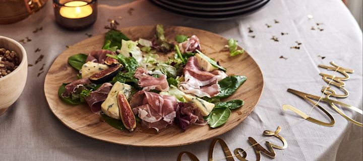 Vi har 4 enkle opskrifter på forretter til din nytårsfest, som du kan prøve. En lækker salat med prosciutto og blåskimmelost ved siden af et godt nytårsskilt.  