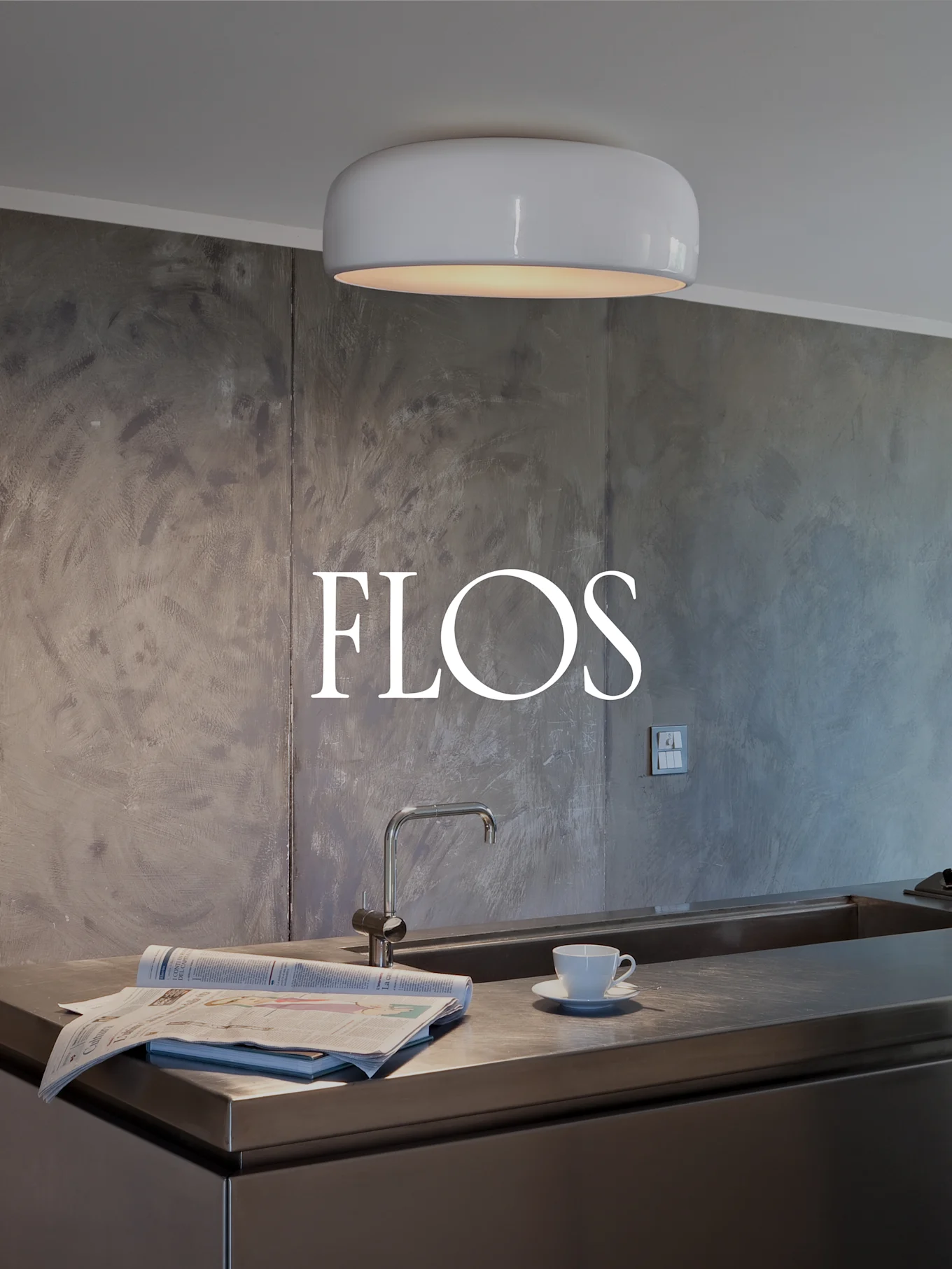 Moderne køkken med hvid, afrundet loftslampe, bordplade i rustfrit stål med vask, avis og tekop. FLOS-logo på tekstureret væg.
