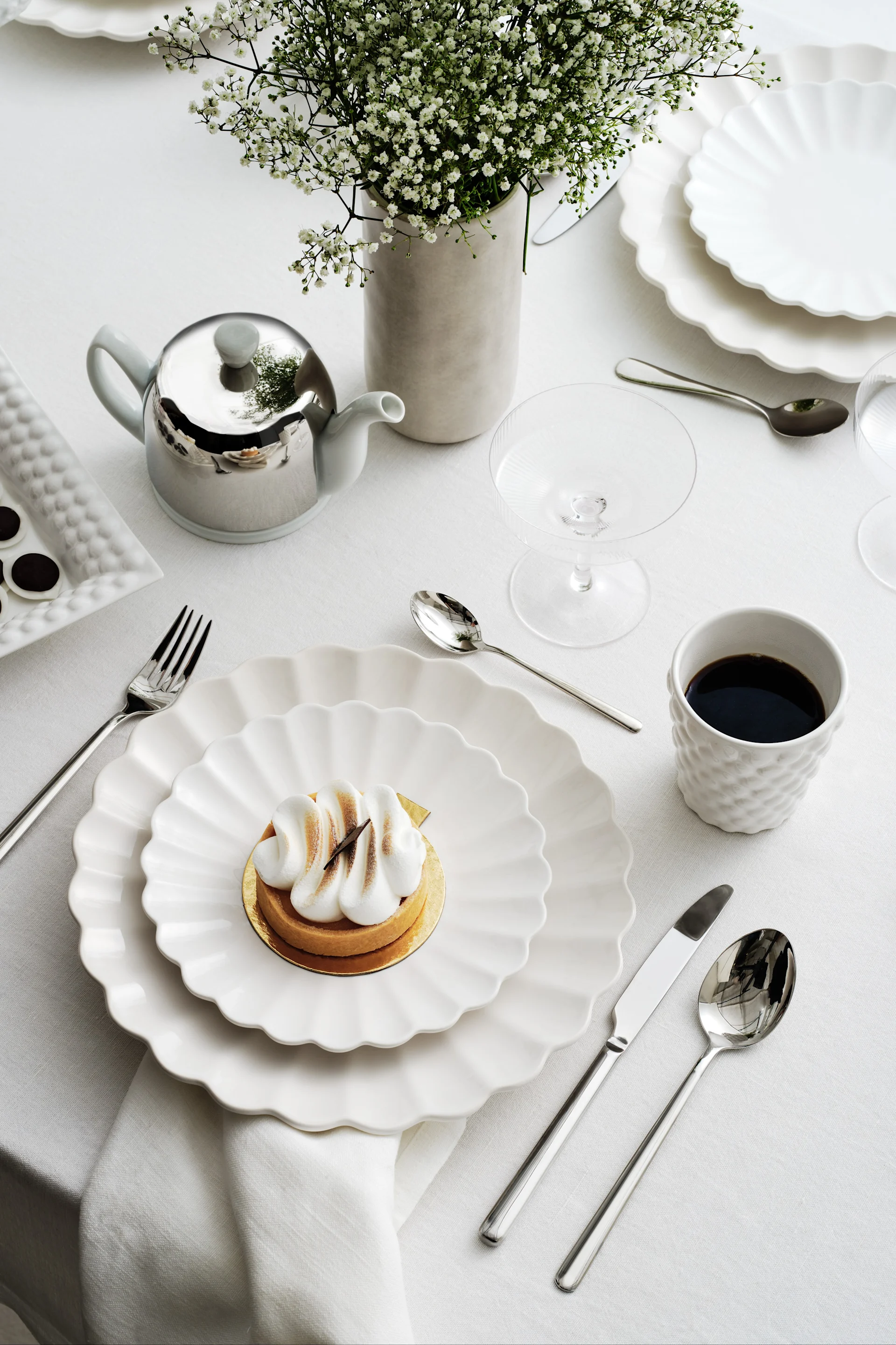 Elegant borddækning med hvide bølgede tallerkener, en marengstærte, kaffe, en sølvtekande og brudeslørblomster.