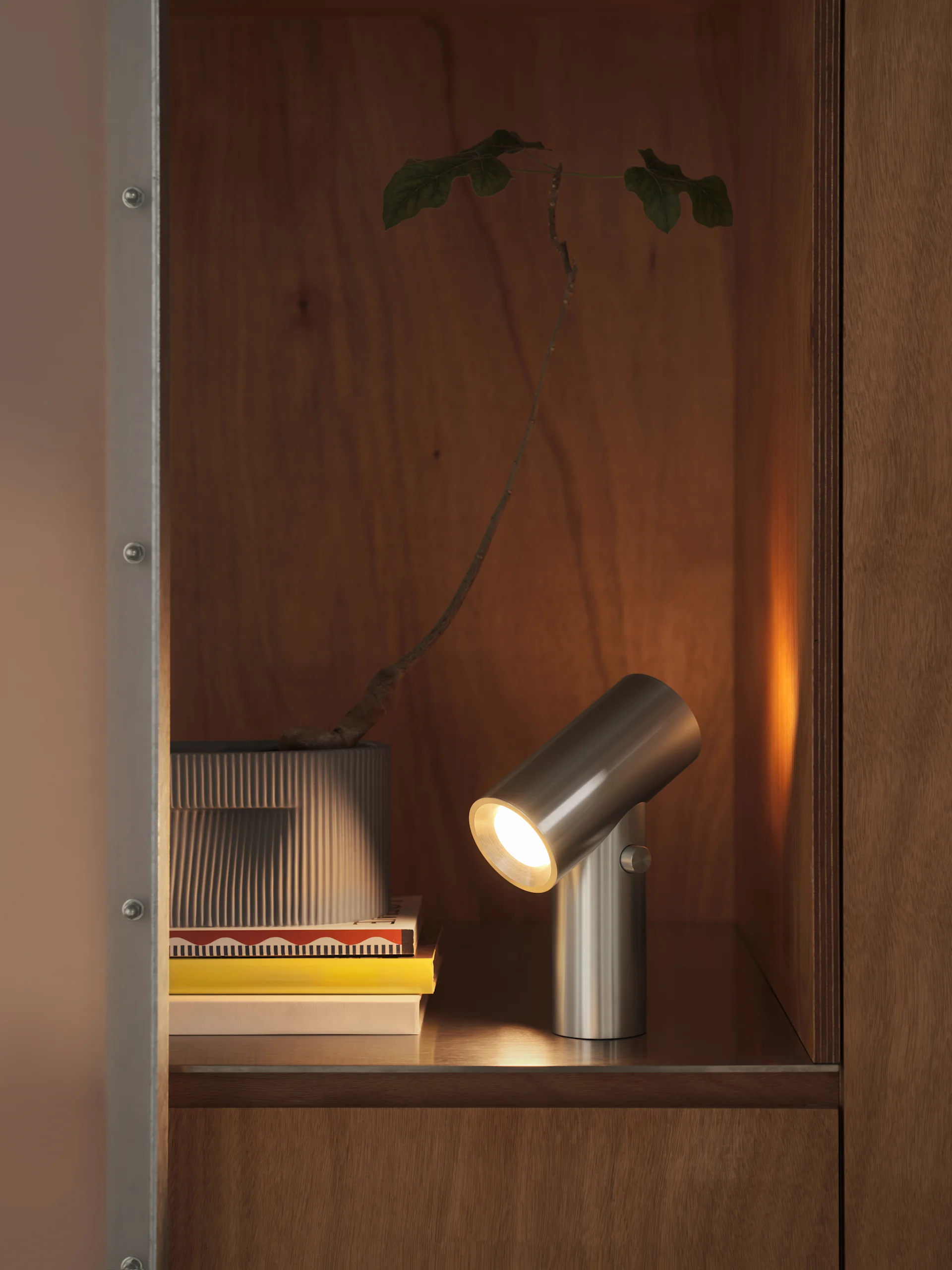 En børstet metal cylindrisk lampe oplyser en stak bøger og en plante i en riflet potte, inde i et træskab.