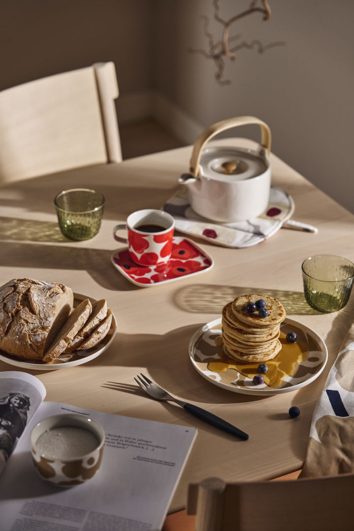 Et morgenmadsbord er dækket med Marimekko Unikko tallerkener og en tekande og en stak pandekager med sirup.