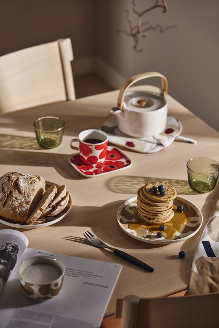 Et morgenmadsbord er dækket med Marimekko Unikko tallerkener og en tekande og en stak pandekager med sirup.