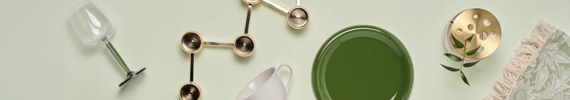 Flad layout: glas med grøn stilk, guldsmykket indretning, hvidt krus, grøn tallerken, messinggenstand med blade og grøn mønstret tekstil med frynser.
