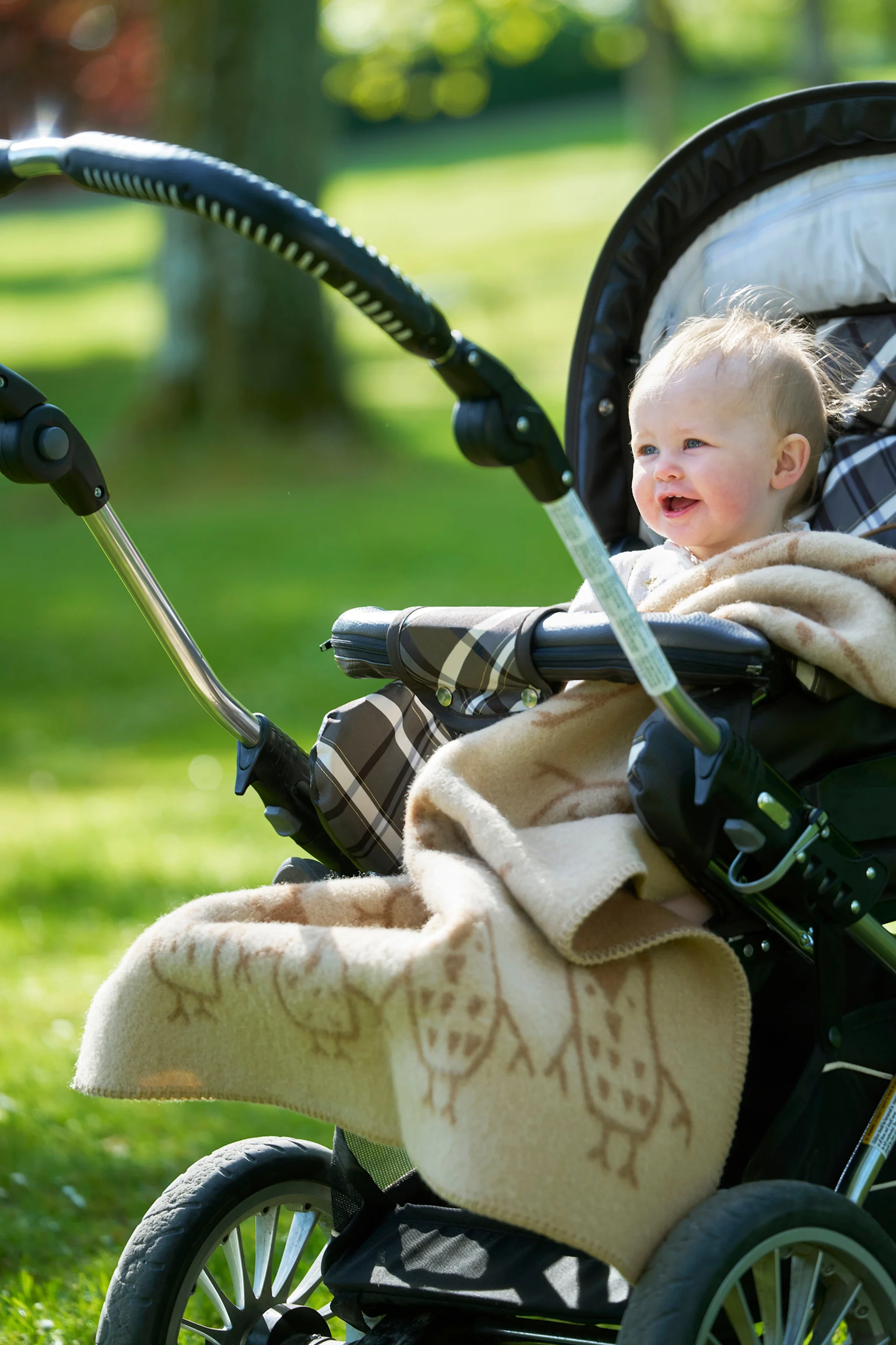 En smilende baby med lyst hår, pakket ind i et tæppe med ugle-mønster, sidder i en barnevogn udendørs i en solrig park.