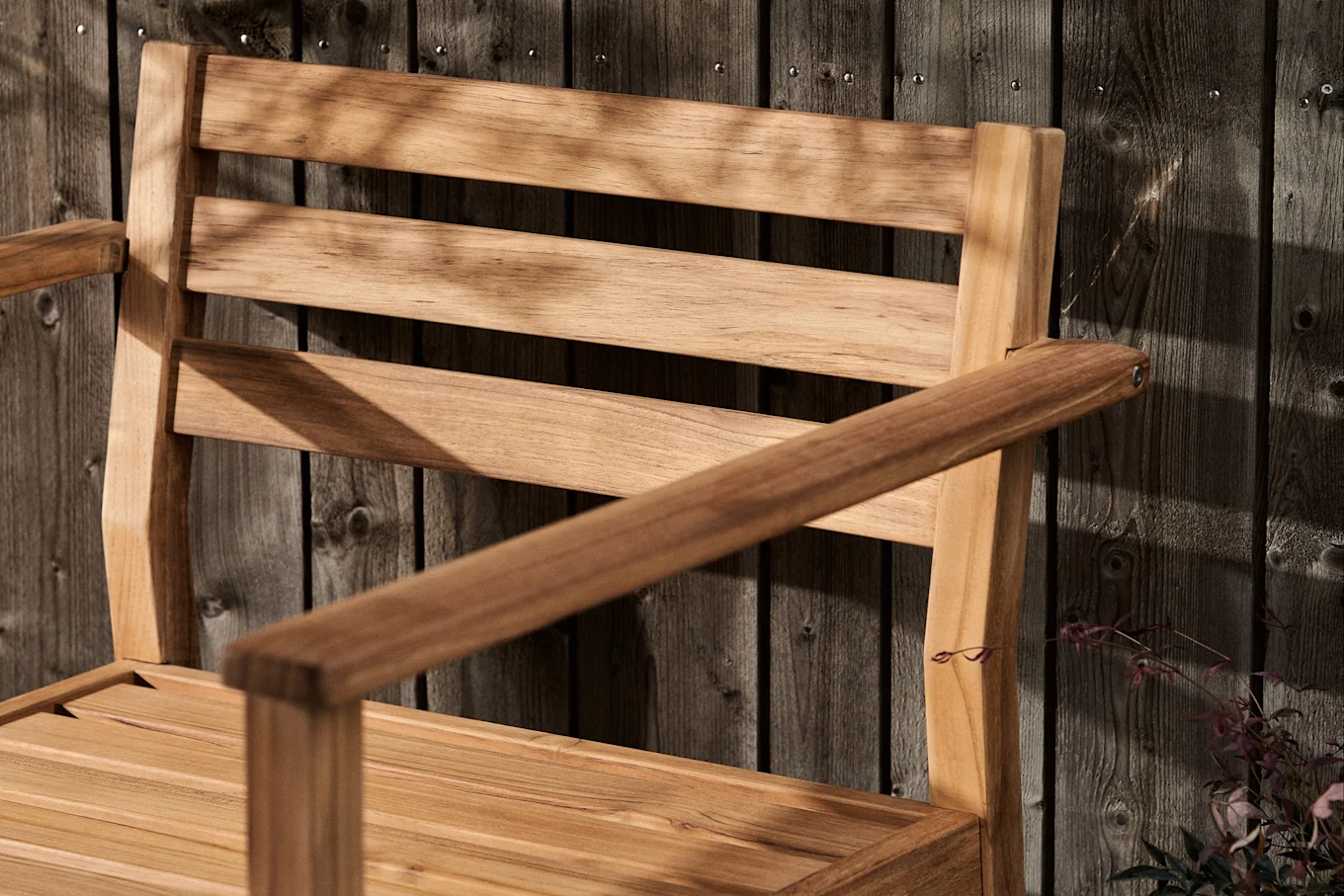 Lamellæggeret træstol med armlæn mod et mørkt træhegn, dækket af bladskygger.