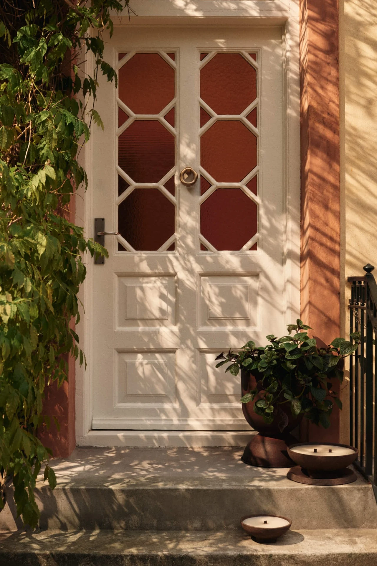 Hvid hoveddør med ottekantede glaspaneler, grønne slyngplanter til venstre og en urtepotte med to stearinlys på trappen.