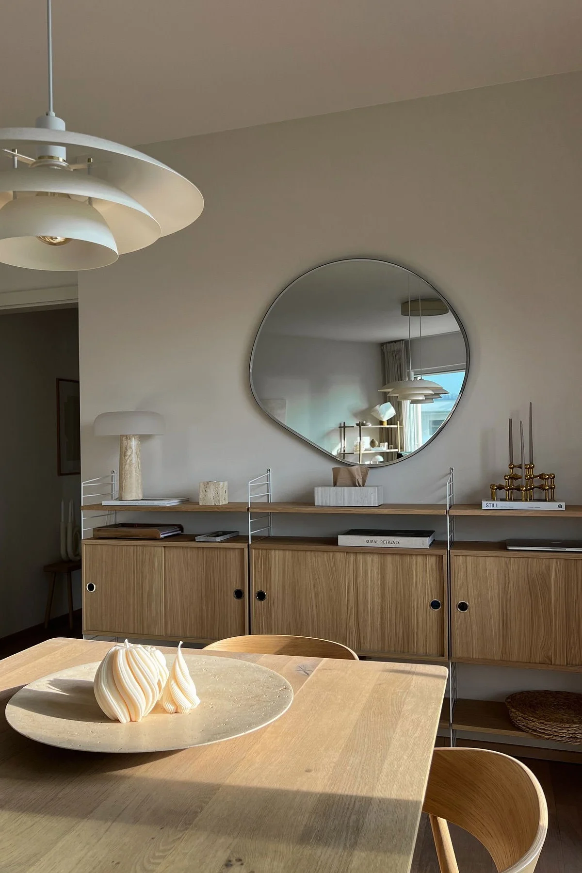 Moderne spisestue med træbord, to hvide spirallys, træhylde, uregelmæssigt spejl og lagdelt lampe.