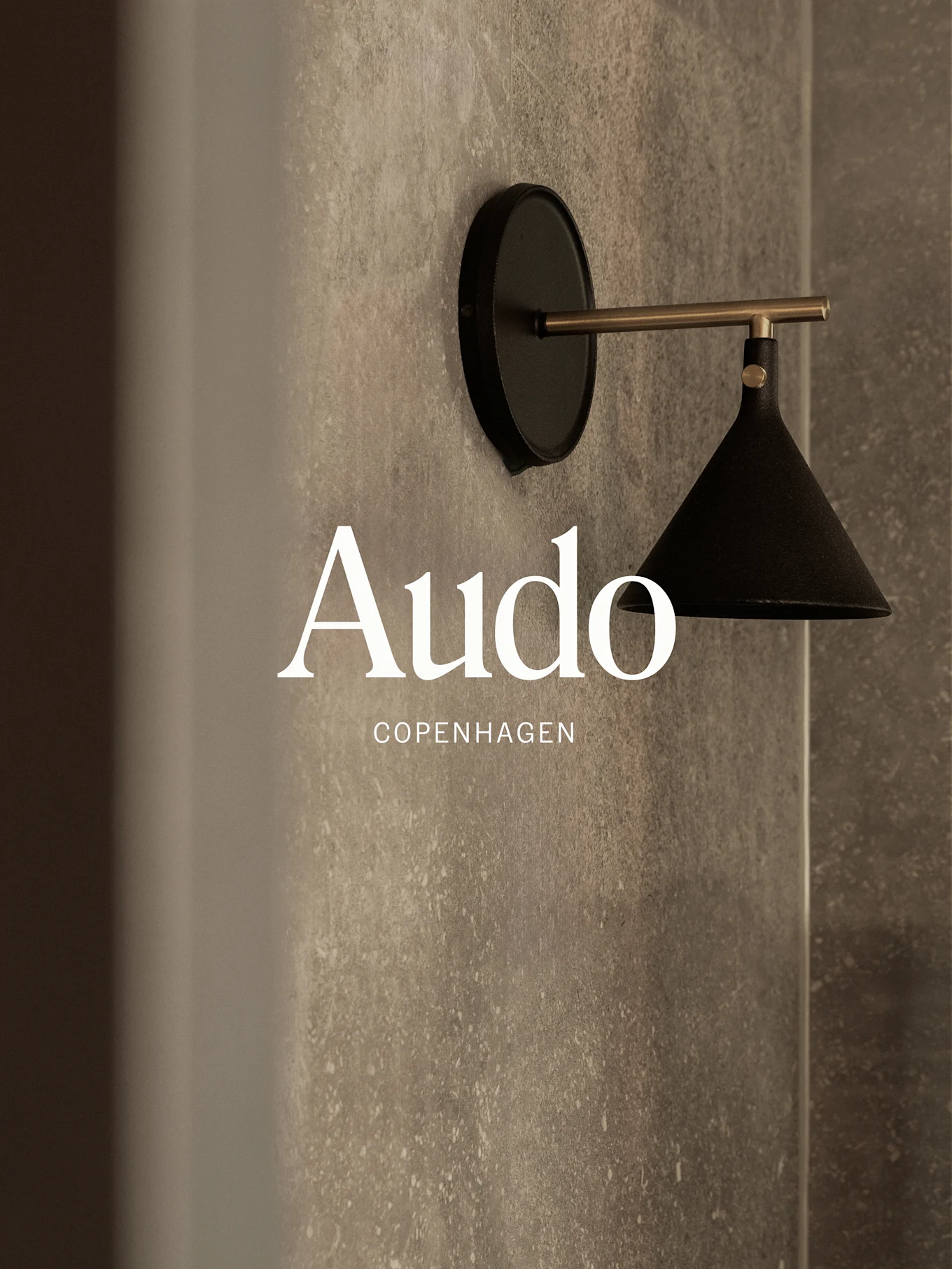 En vægmonteret sort lampe med en messingarm mod en tekstureret grå væg, med teksten 'Audo Copenhagen'.