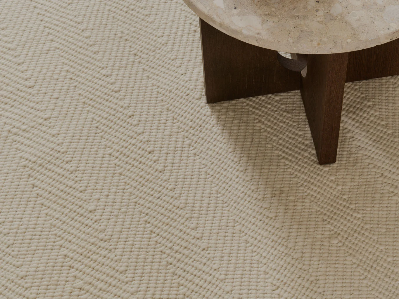 Nærbillede af et cremefarvet tekstureret tæppe med sildebensmønster, ved siden af et bord med stenplade.