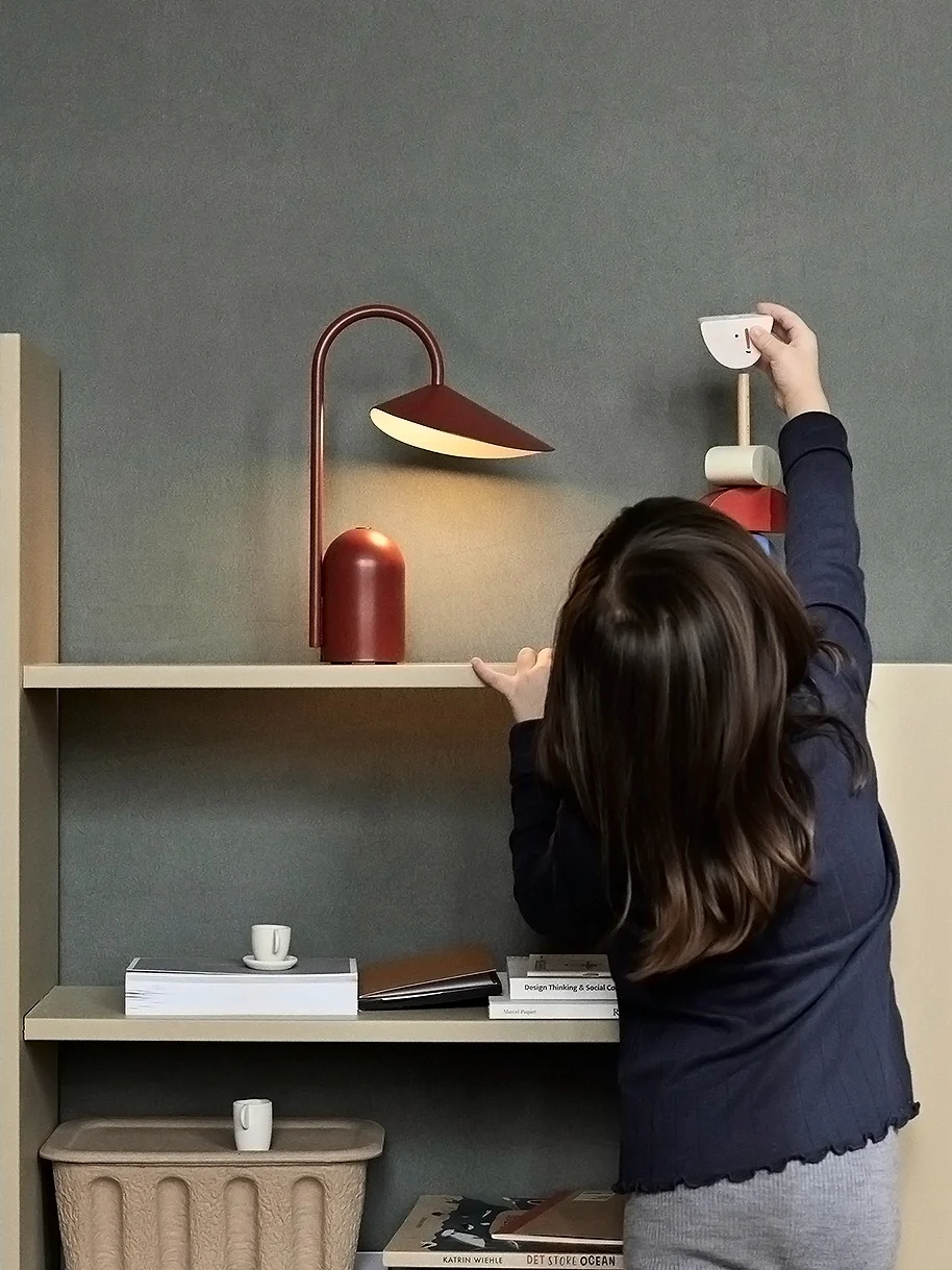 Et barn rækker ud efter et træfuglelegetøj på en lys hylde med en rød lampe og bøger; en flettet kurv står på gulvet.