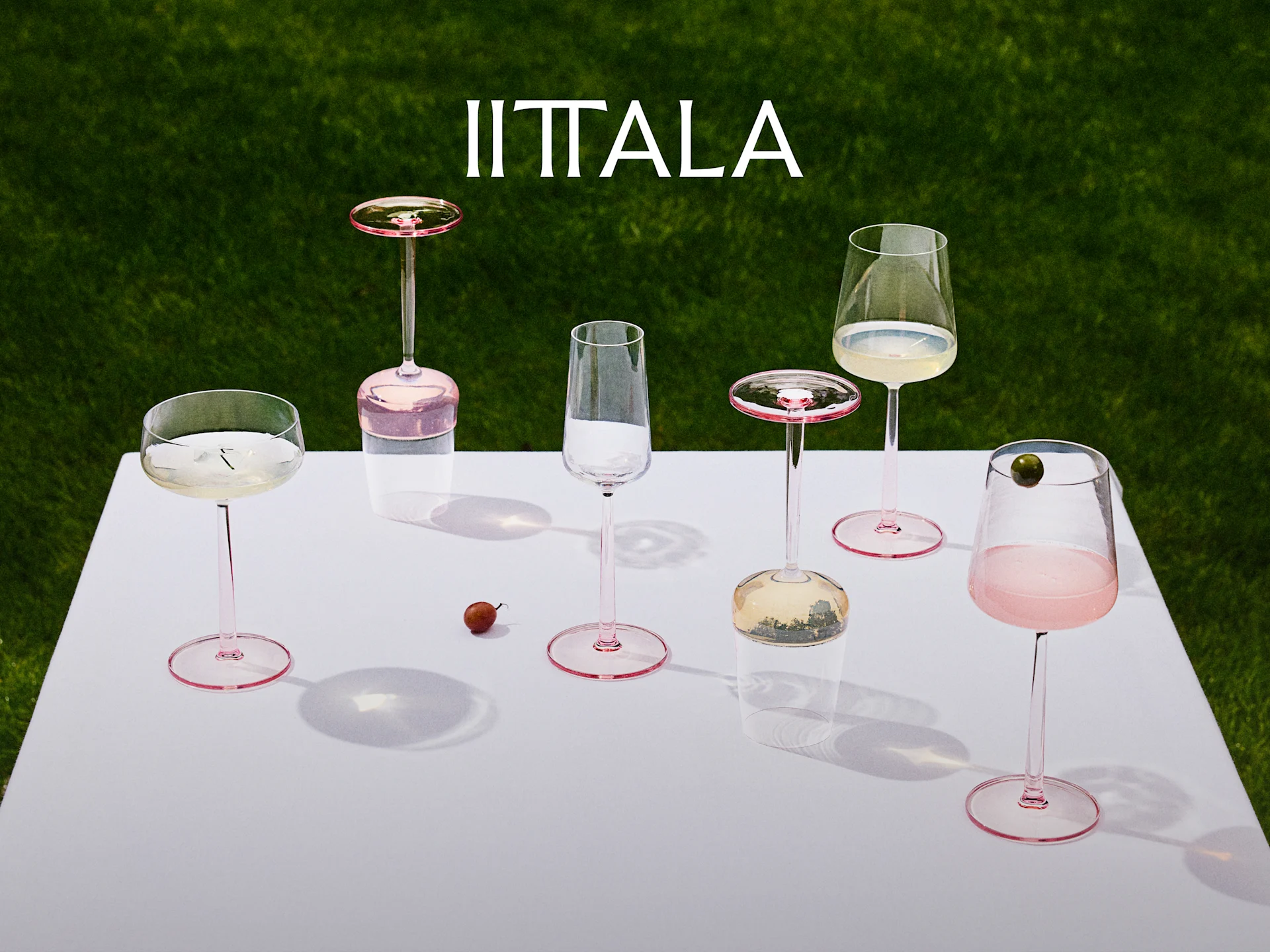 Iittala-logo over en samling lyserøde vinglas på et hvidt bord med grønt græs i baggrunden, nogle med drinks.