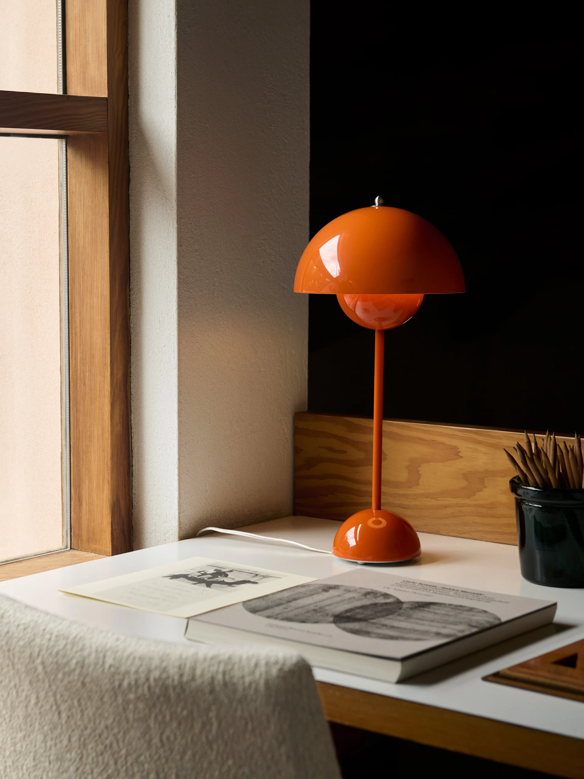 En orange, kuppelformet bordlampe står på et hvidt skrivebord ved siden af bøger og en beholder med blyanter.