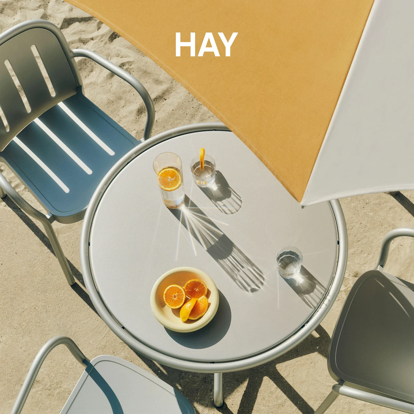 Oversigt over et rundt gråt udebord med to stole på sand, med drikkevarer og appelsiner under en gul og hvid parasol med 'HAY'.