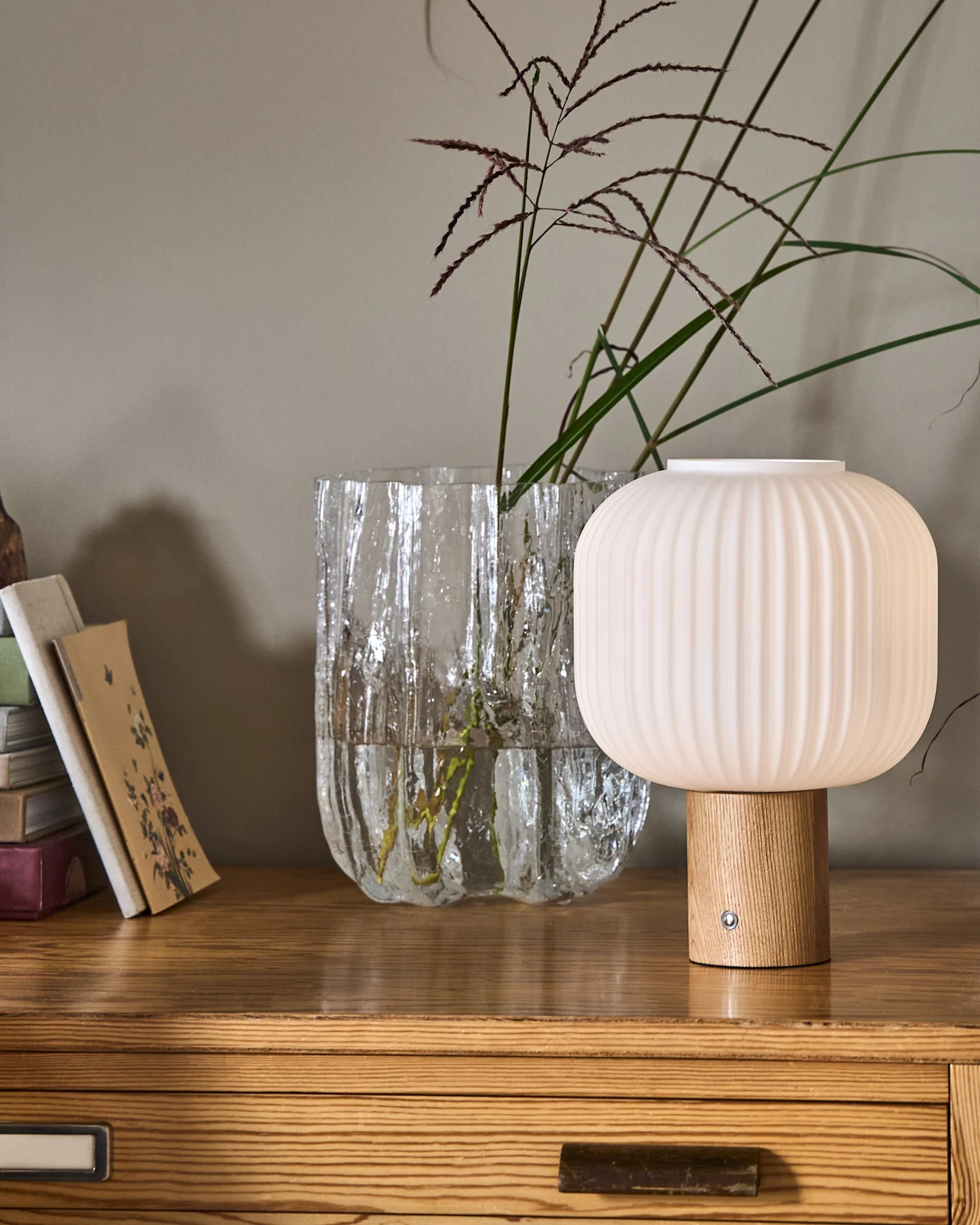 En rillet, champignonformet lampe med træfod ved siden af en tekstureret glasvase med planter og en stak bøger på en trækommode.