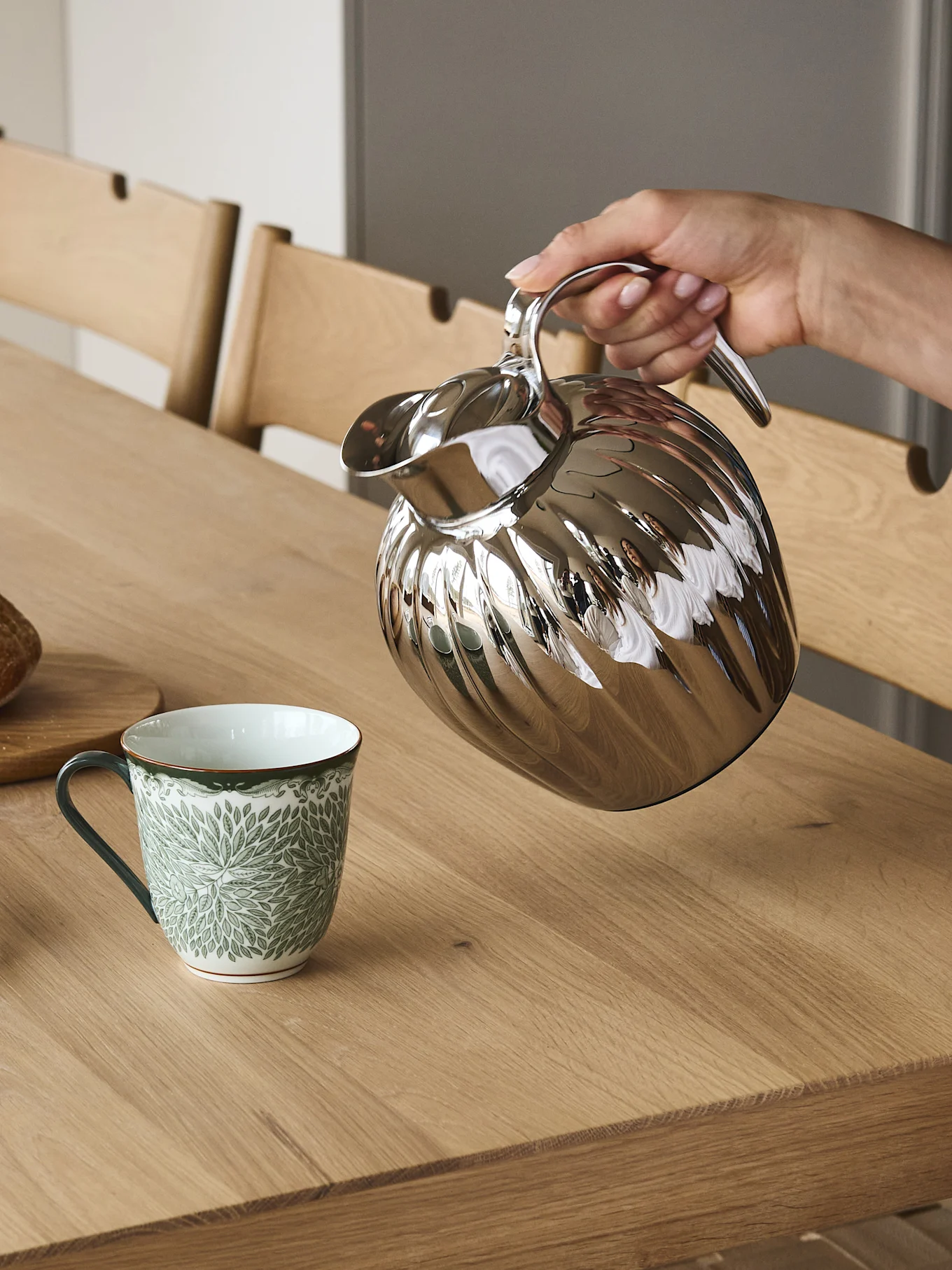 En hånd hælder fra en sølvfarvet riflet termokande ned i et grønt og hvidt mønstret krus på et træbord.