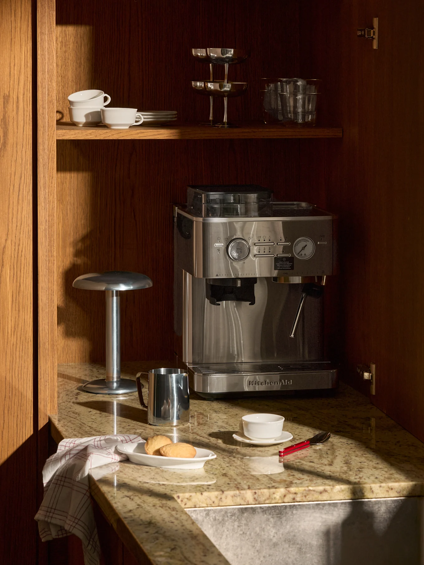 En espressomaskine i rustfrit stål på granitbord i træskab. Ovenover kopper og glas. På bordet småkager, kop og mælkekande.