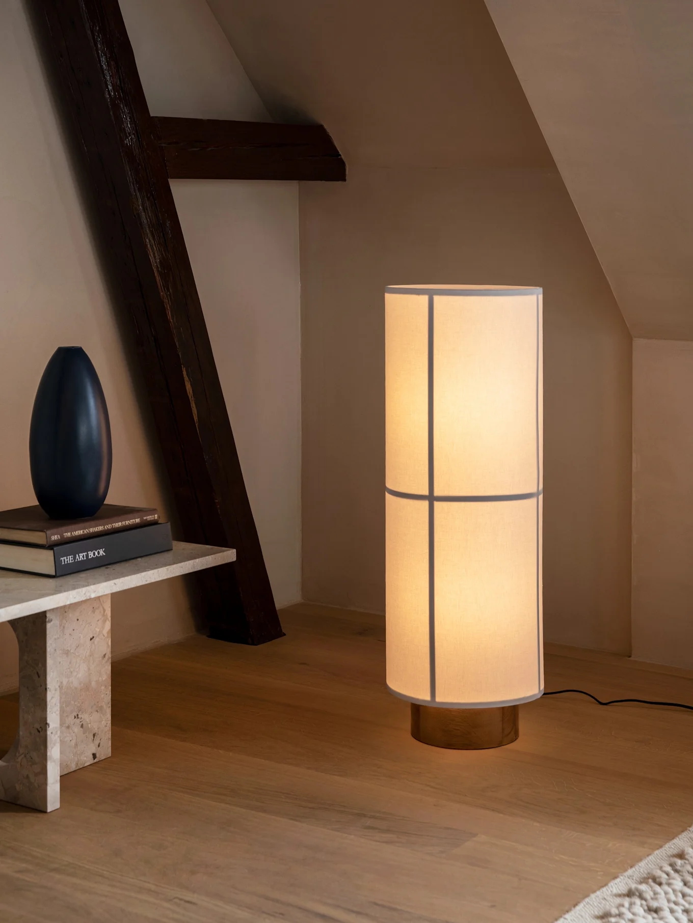 Høj cylindrisk gulvlampe, lys skærm og træfod, oplyser rum med stenbænk og mørk bjælke.