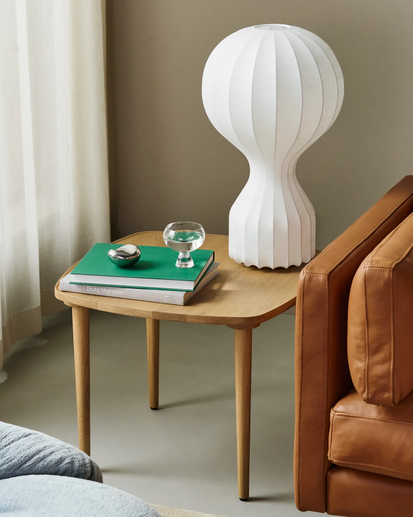 Trækonsolbord med hvid riflet lampe, grøn bog, vandglas, sølvdekoration ved brun lædersofa.