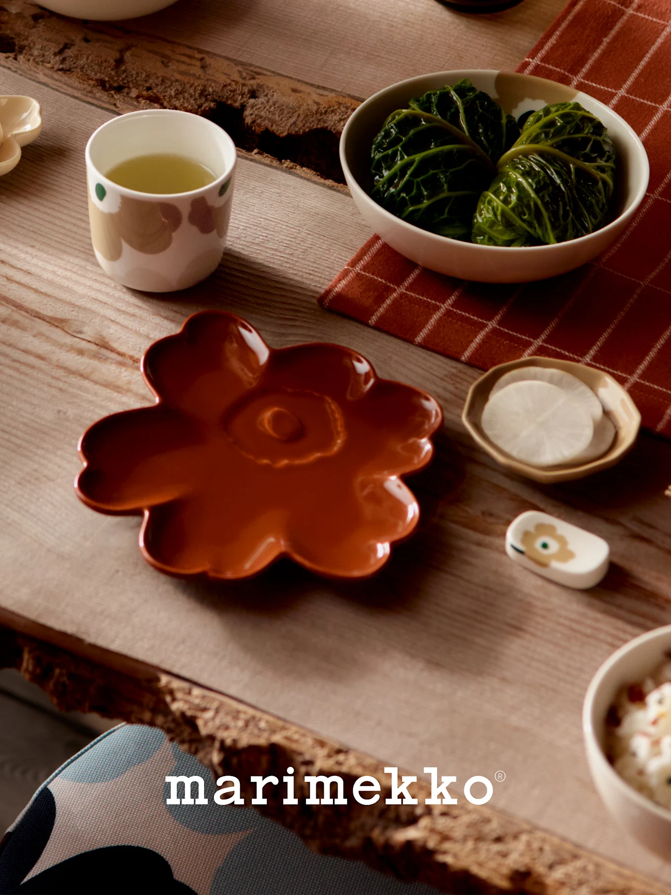 Bord dækket med Marimekko-service: en rustfarvet blomstertallerken, en kop grøn te, kålruller og skiveskårne radiser.