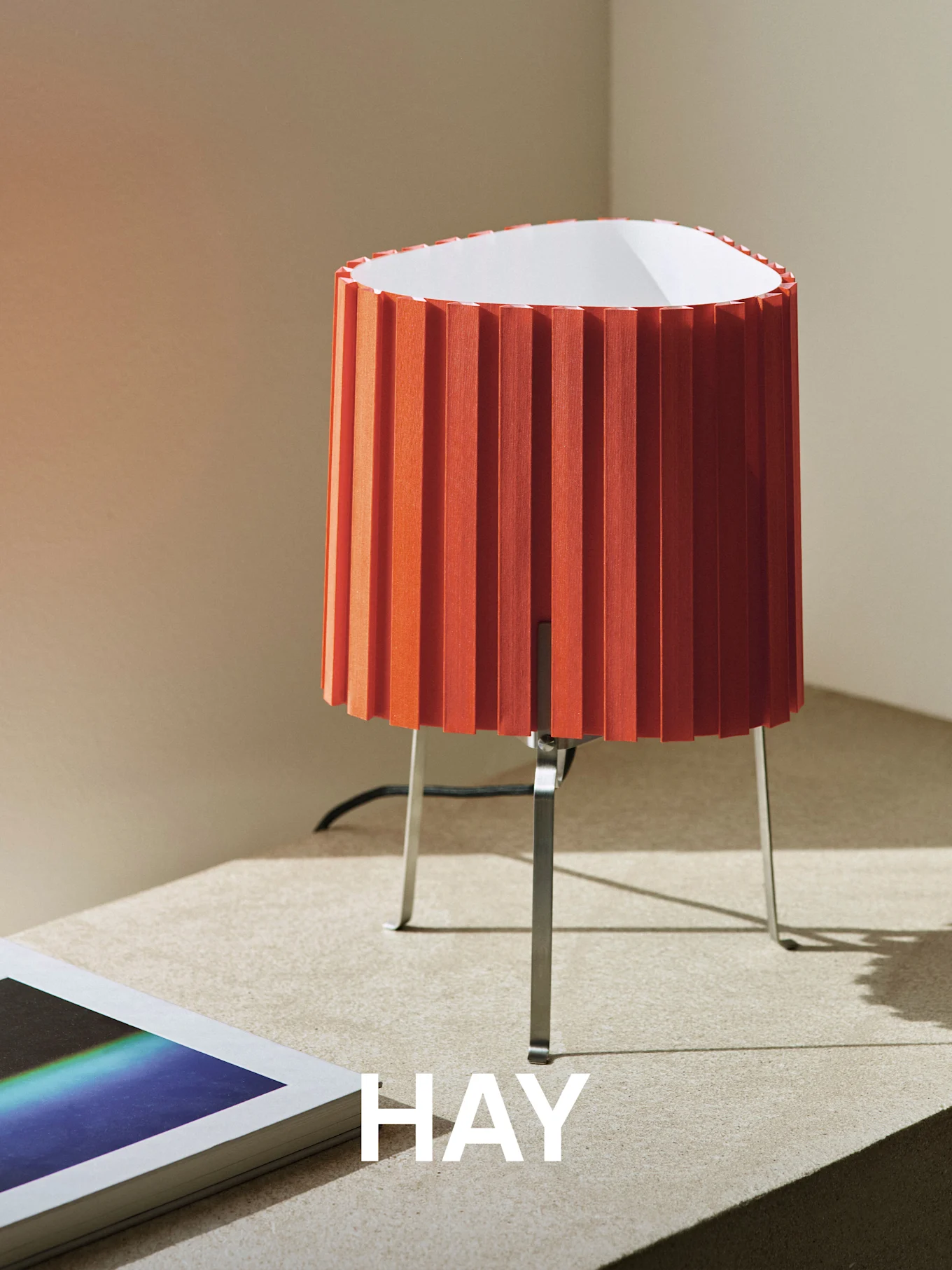 Orange plisseret bordlampe med metalfod på en tekstureret overflade, ved siden af en bog. Ordet 'HAY' er overlejret.