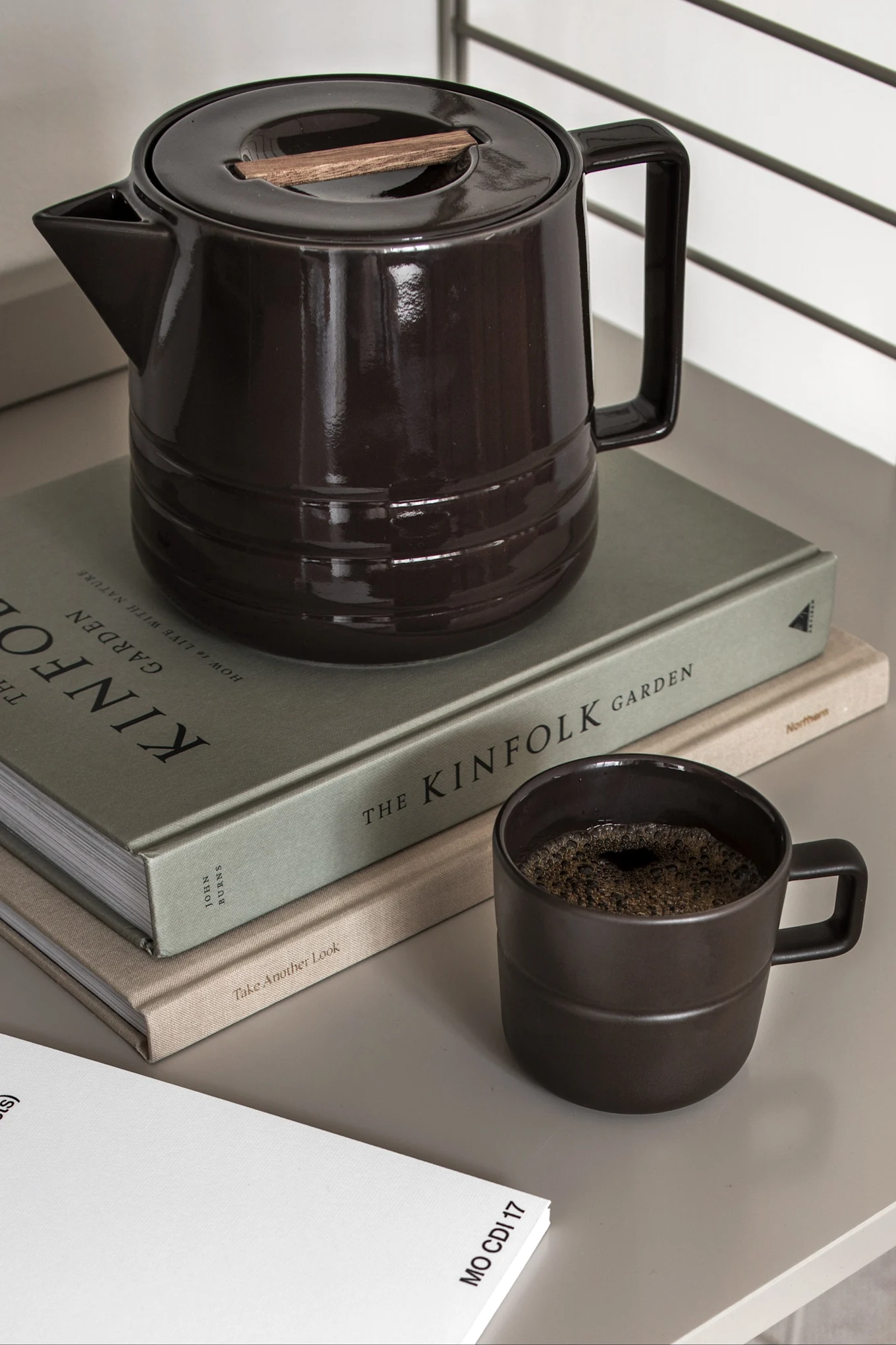 Mørkebrun keramisk kaffekande med trælåg, matchende krus med kaffe og to bøger på en lys overflade.