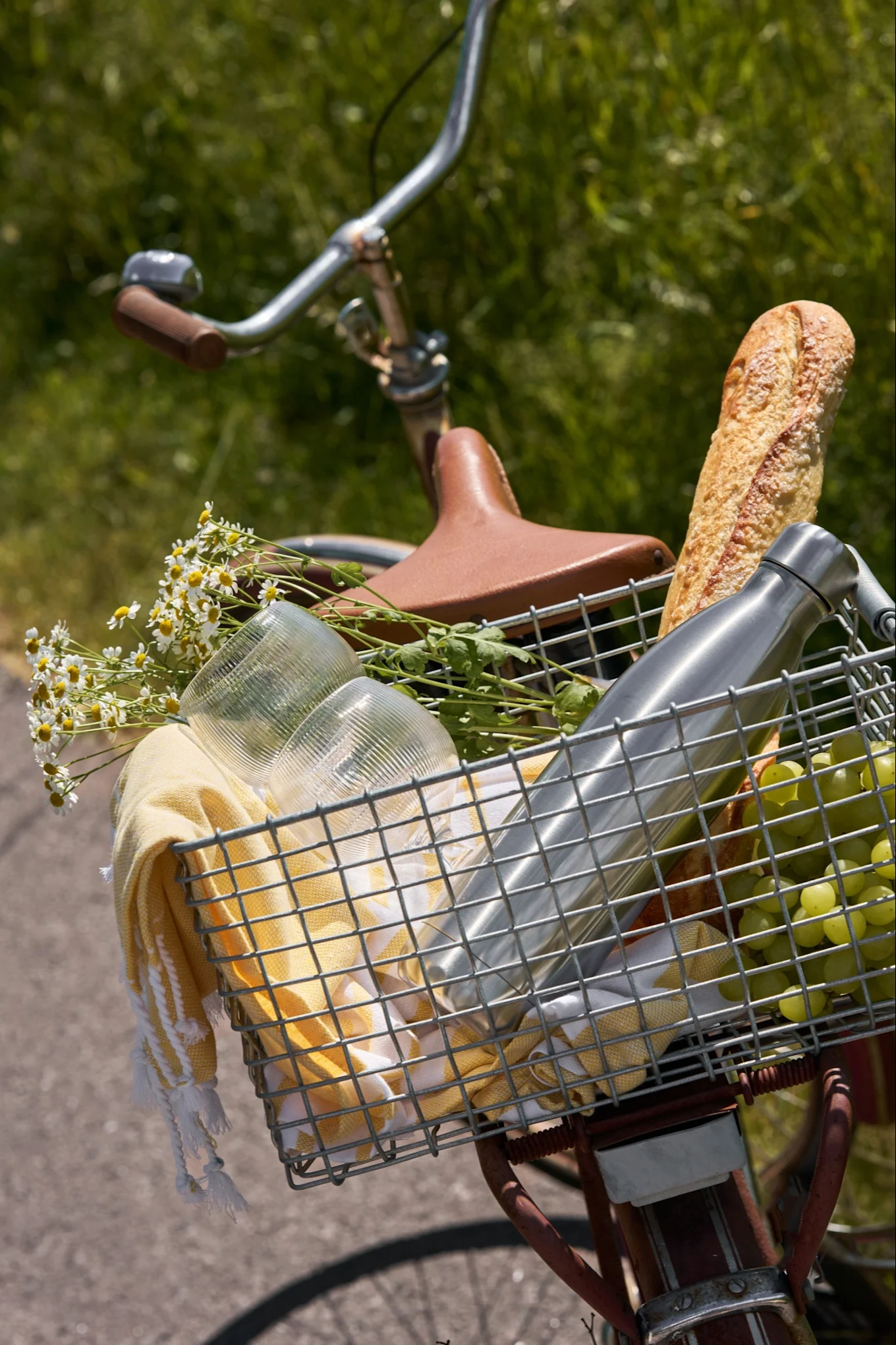 En cykels forkurv fyldt med picnicting: baguette, vandflaske, druer, kopper, gult håndklæde og hvide margeritter.