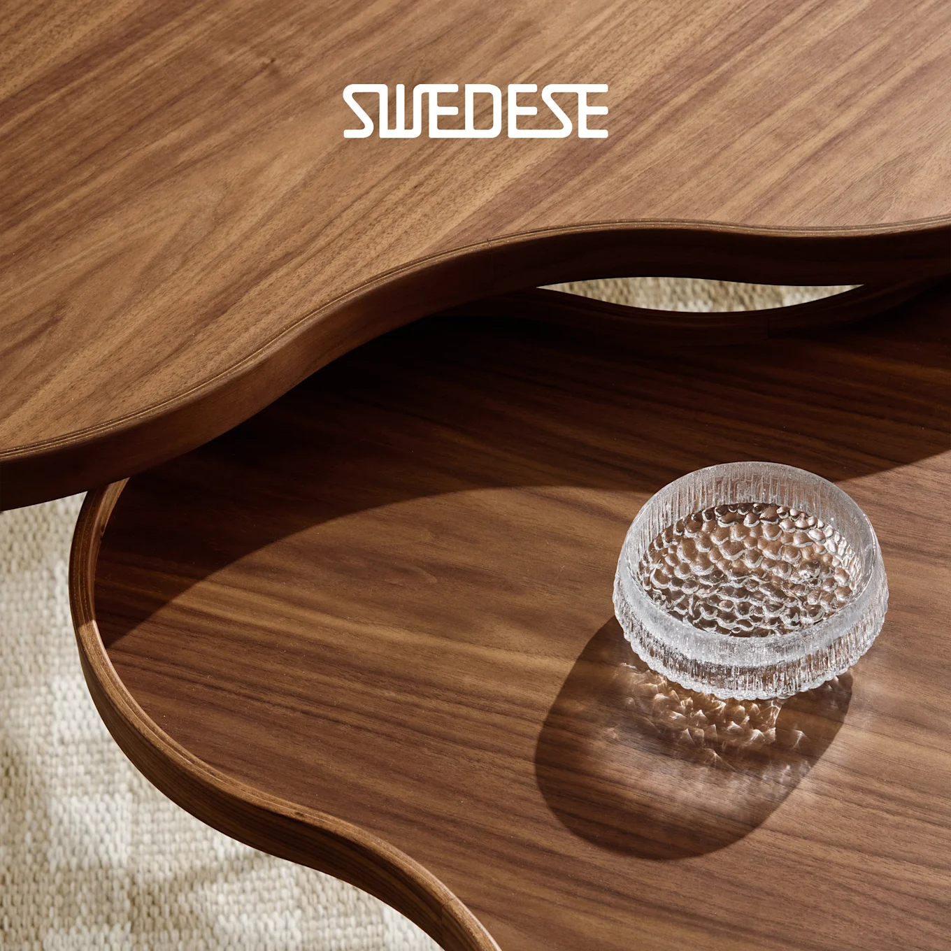 To buede valnøddetræsborde, et overlappende det andet, med en tekstureret klar glasskål på det nederste bord. SWEDESE logo.