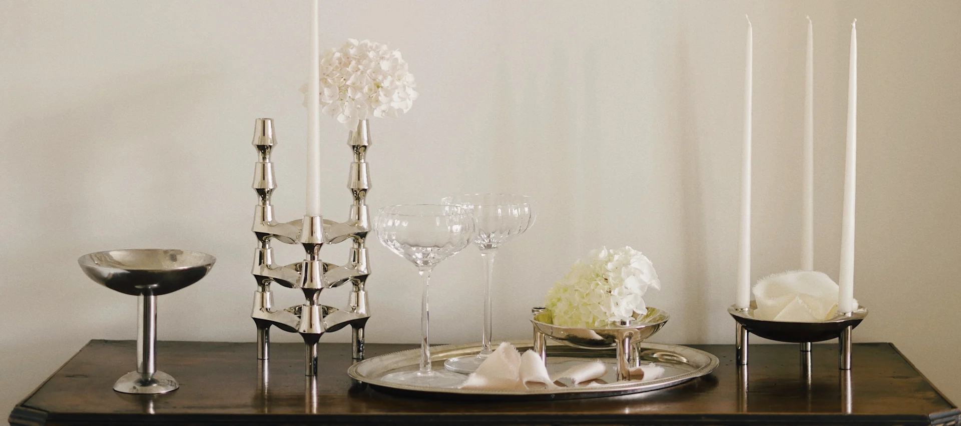 Elegant borddekoration med sølvstager, hvide lys, glaskopper og hvide hortensia på træoverflade.