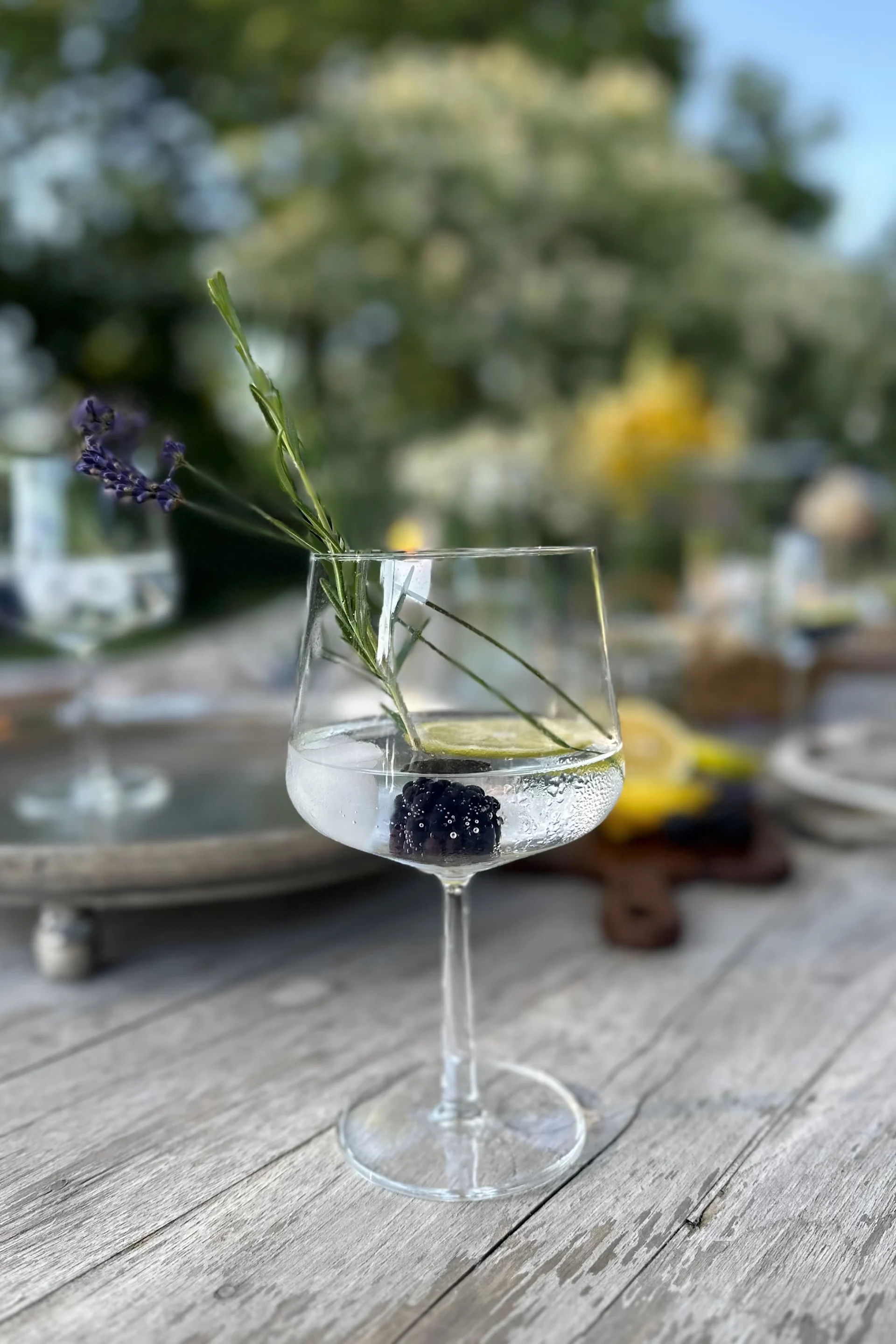En cocktail i et stilkglas med et brombær, limeskive, lavendel og en grøn urt på et træbord.