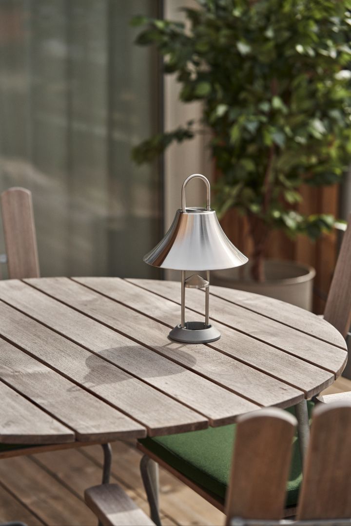 Skab en god atmosfære i din have eller terrasse med Mousqueton portabel lampe i rustfrit stål fra HAY, stående på et rundt teak udendørsbord.