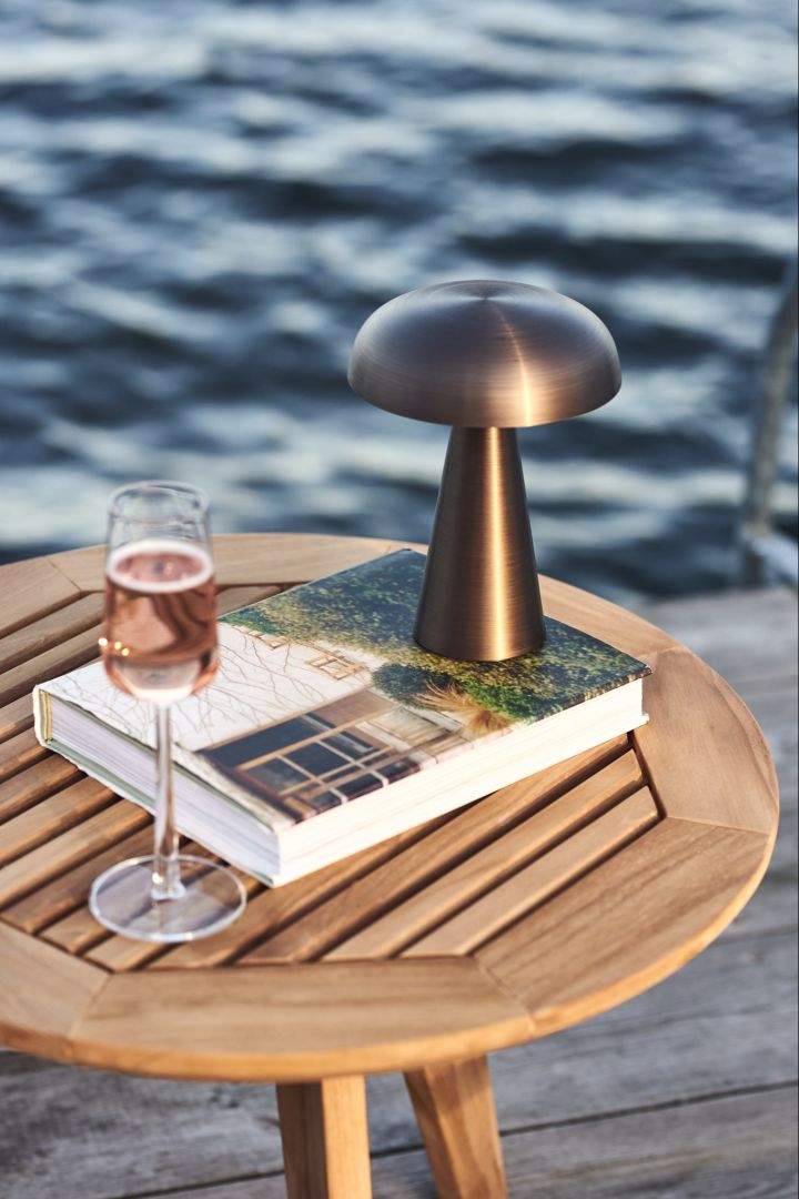 Den trådløse lampe, Como fra &Tradition set her på et træbord med et glas vin ved havet.