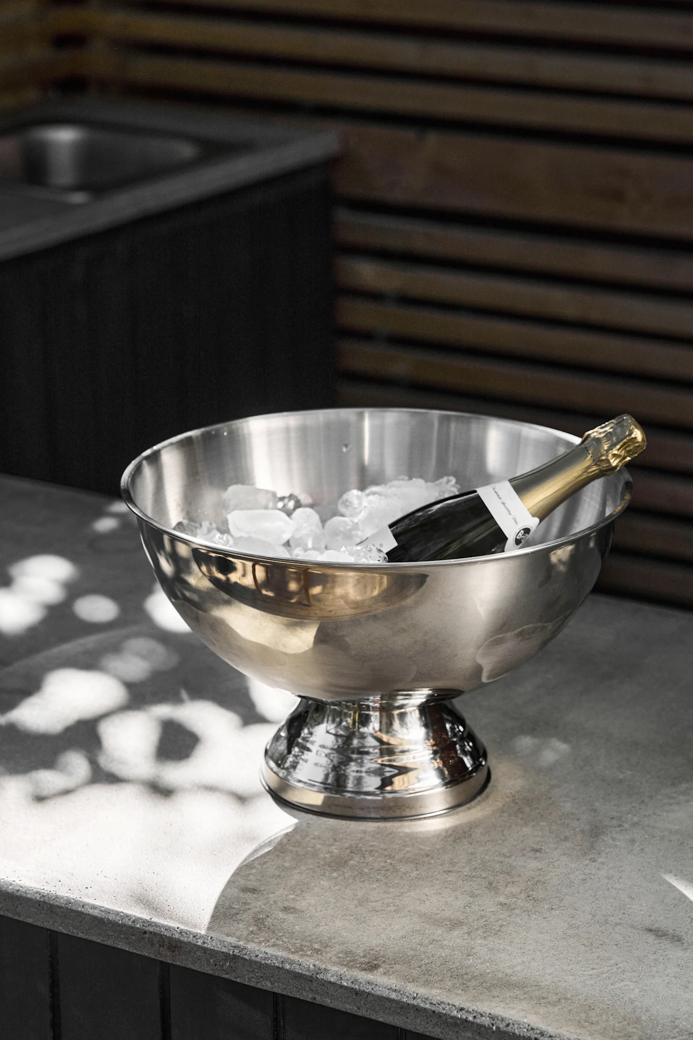 En champagneflaske ligger i en rustfri stålisspand fyldt med isterninger på en grå betondisk, med pletter af sollys.