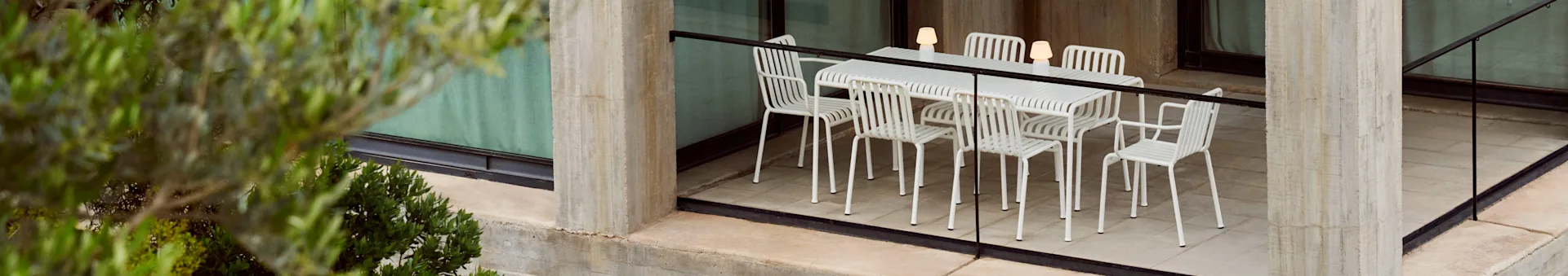 Et udendørs spisesæt med et hvidt lamelbord og seks matchende stole, oplyst af tre bærbare lamper, på en betonterrasse med planter.