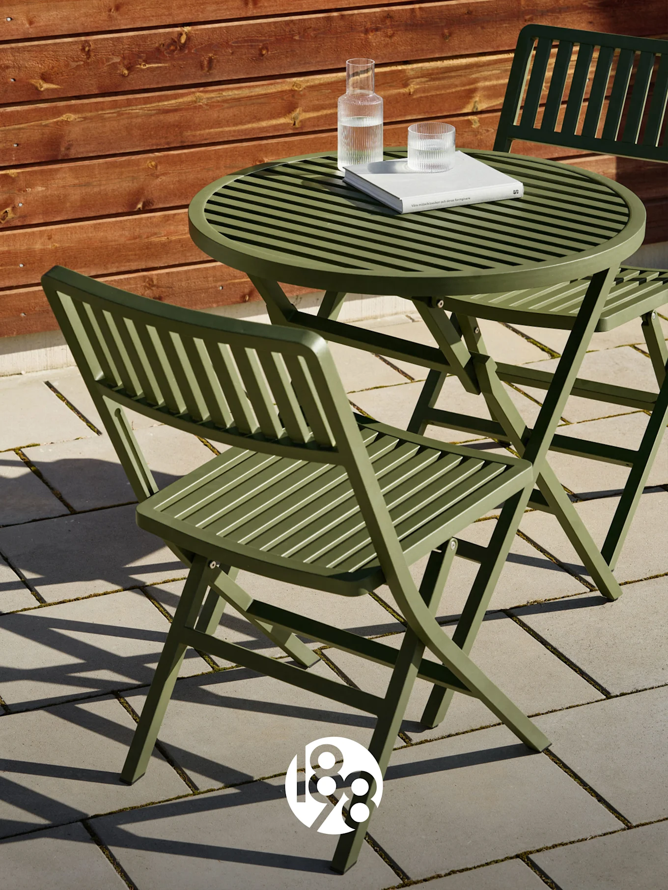 To grønne foldestole med lameller og et matchende bord på en terrasse med en bog og vandglas.