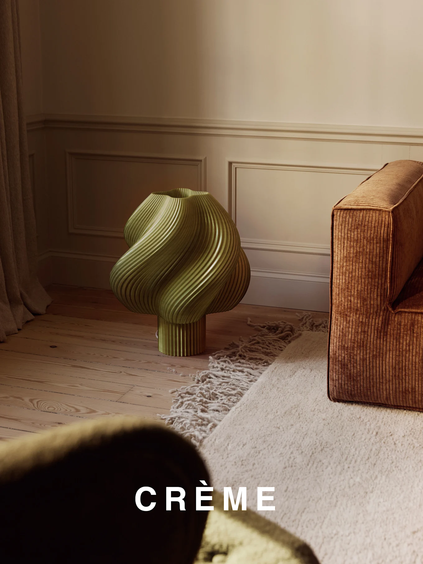 En lysegrøn, snoet ribbet lampe på et trægulv, ved siden af en brun ribbet sofa og et beige langhåret tæppe.