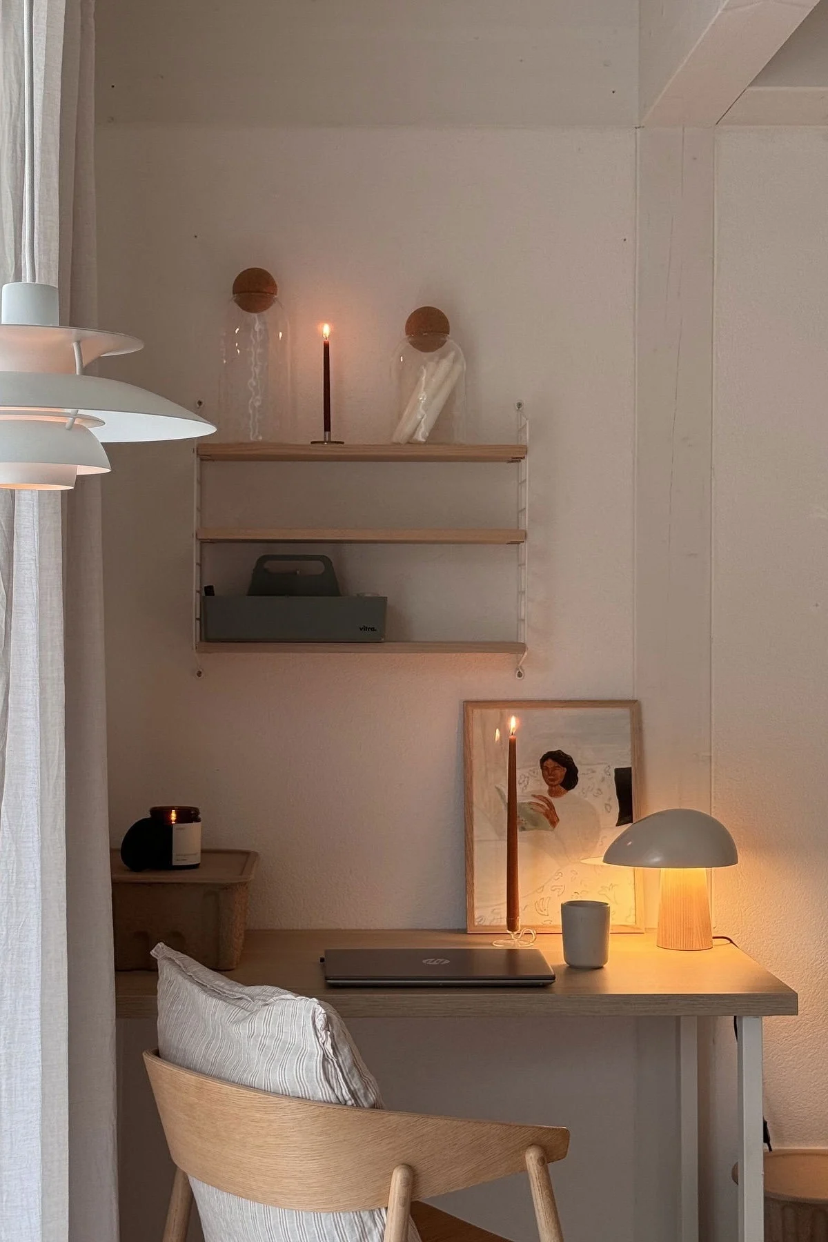 Et hyggeligt skrivebord med varm belysning, en lukket bærbar, to tændte stearinlys, en moderne lampe og et indrammet kunstværk.