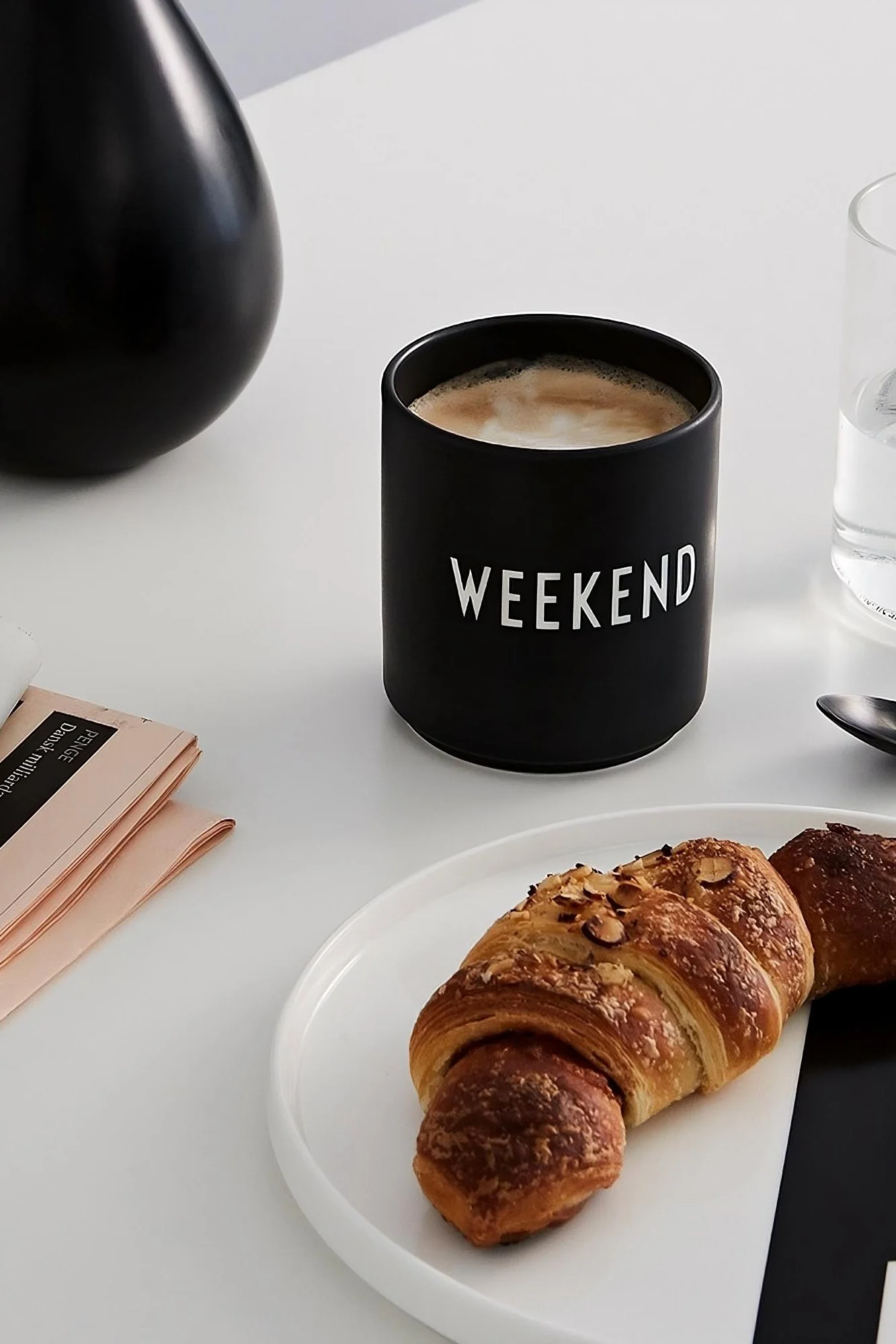 Kaffekrus: Det sorte kaffekrus med Design Letters "Weekend"-printet står på et bord, foran det står en tallerken med en croissant.