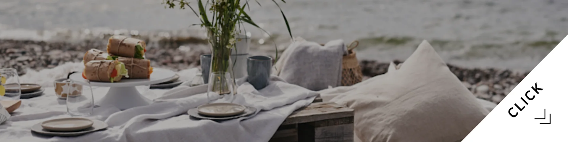 En rolig strandpicnic med indpakket mad, service, en vase med blomster og puder på en stenstrand ved havet.