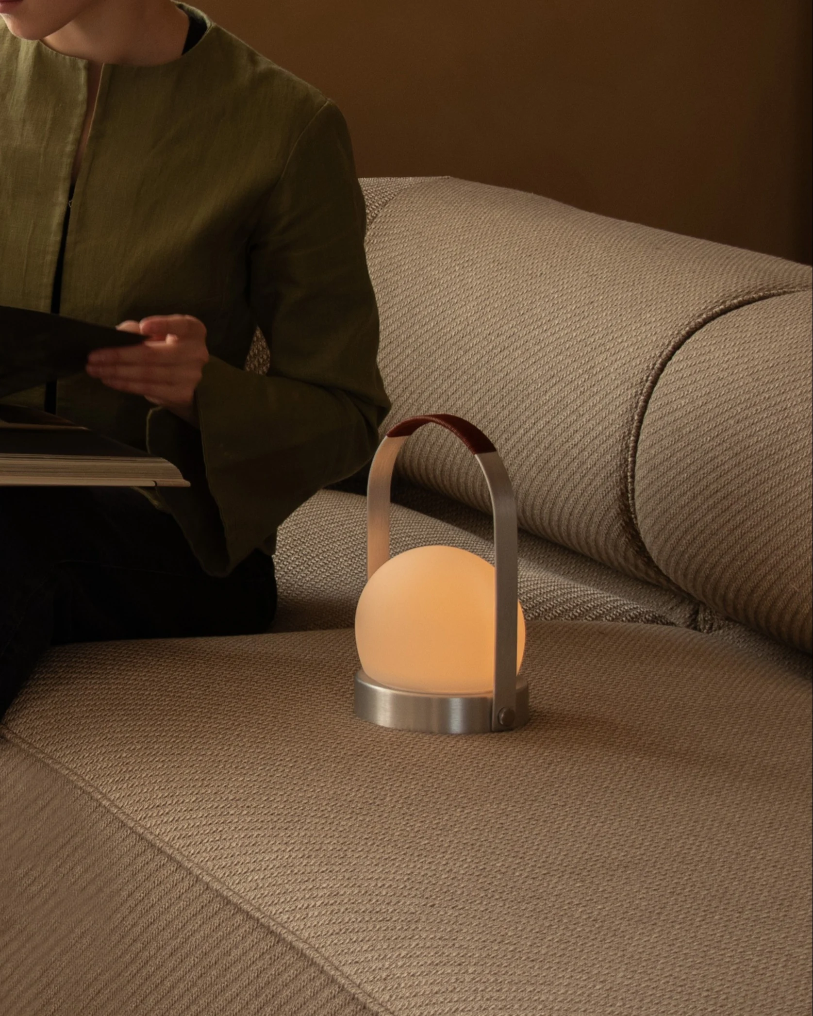 En person læser en bog på en beige sofa ved siden af en glødende kugleformet bærbar lampe med et læderhåndtag.