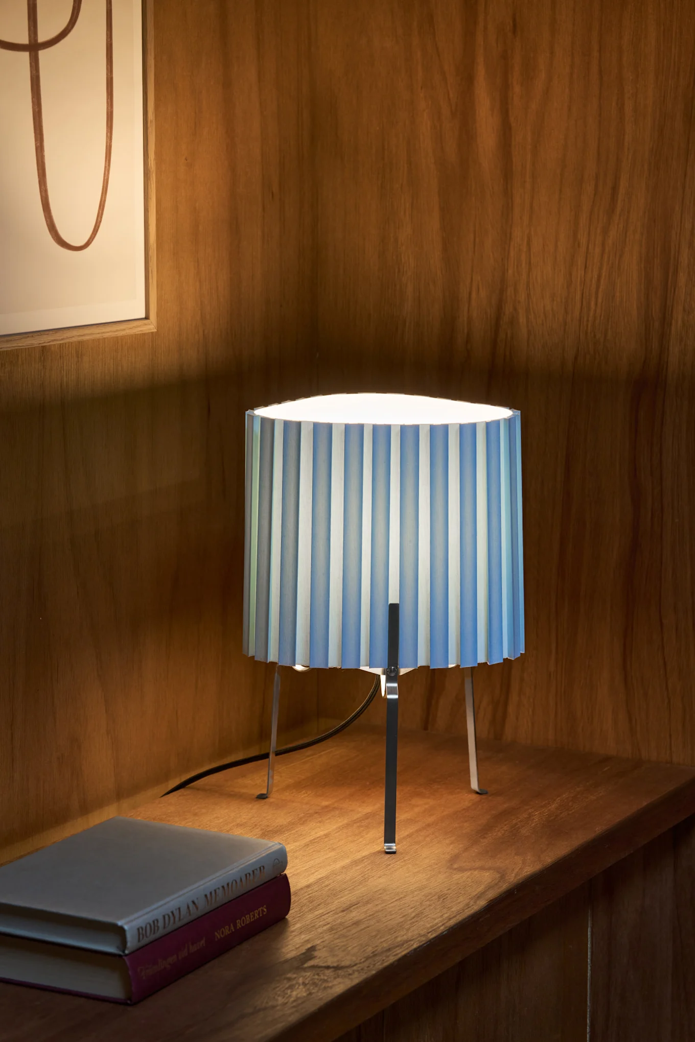 En bordlampe med en blå- og hvidstribet plisseret skærm og metalben, der står på en træhylde ved siden af to bøger.
